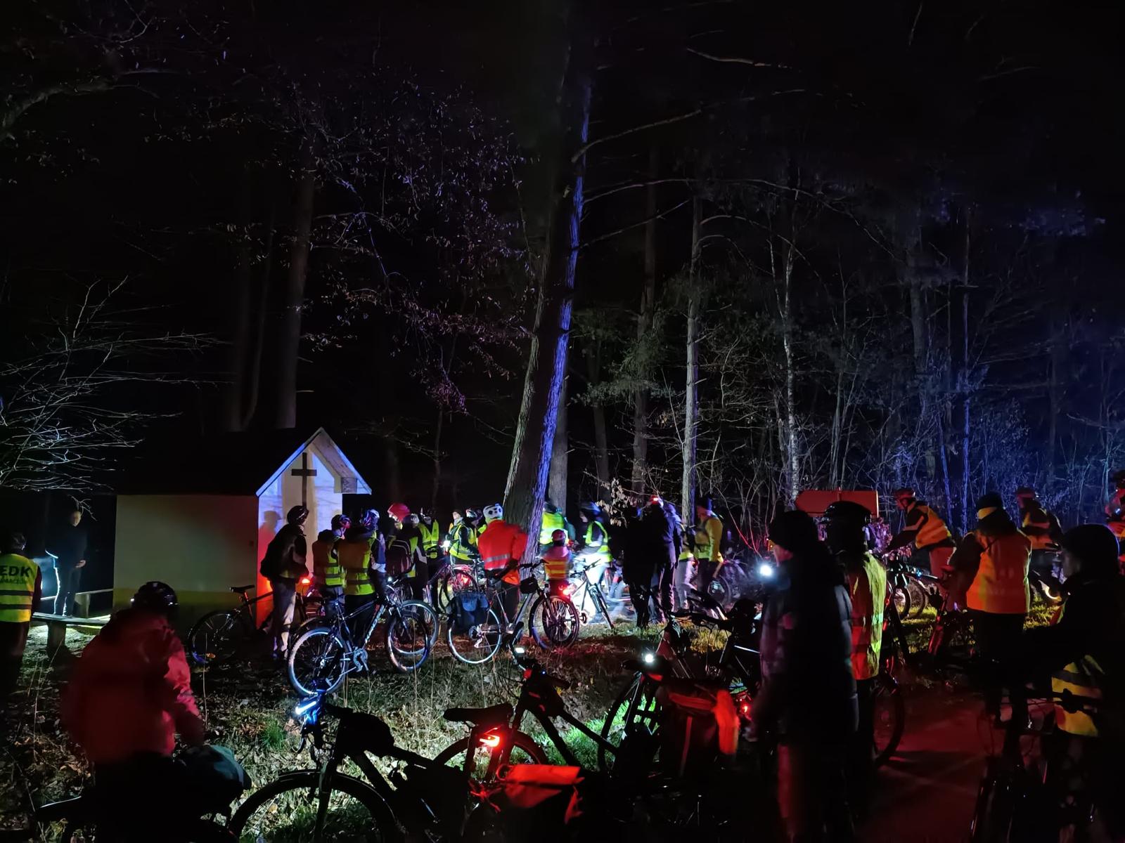 Rowerowa Droga Krzyżowa | Bardzo duża grupa kolarzy stoi w lesie obok murowanej, małej kapliczki. Zdjęcie nocne.jpg
