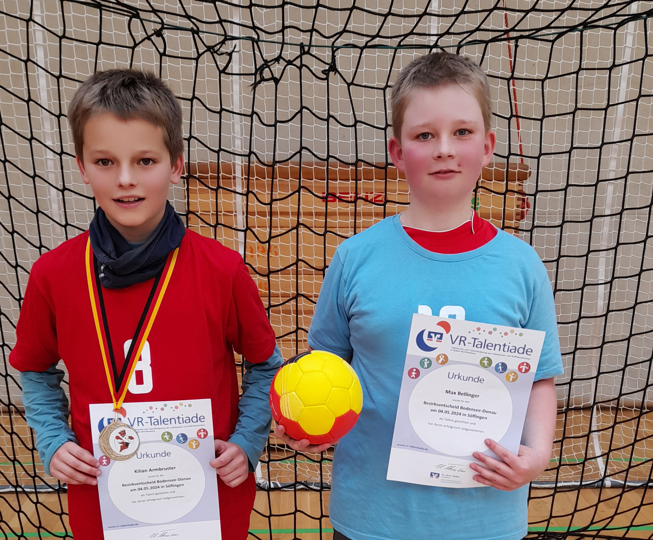 Die zwei Handball-Nachwuchstalente zeigen ihre Urkunden. Den Hintergrund bilden Netze von Handballtoren.