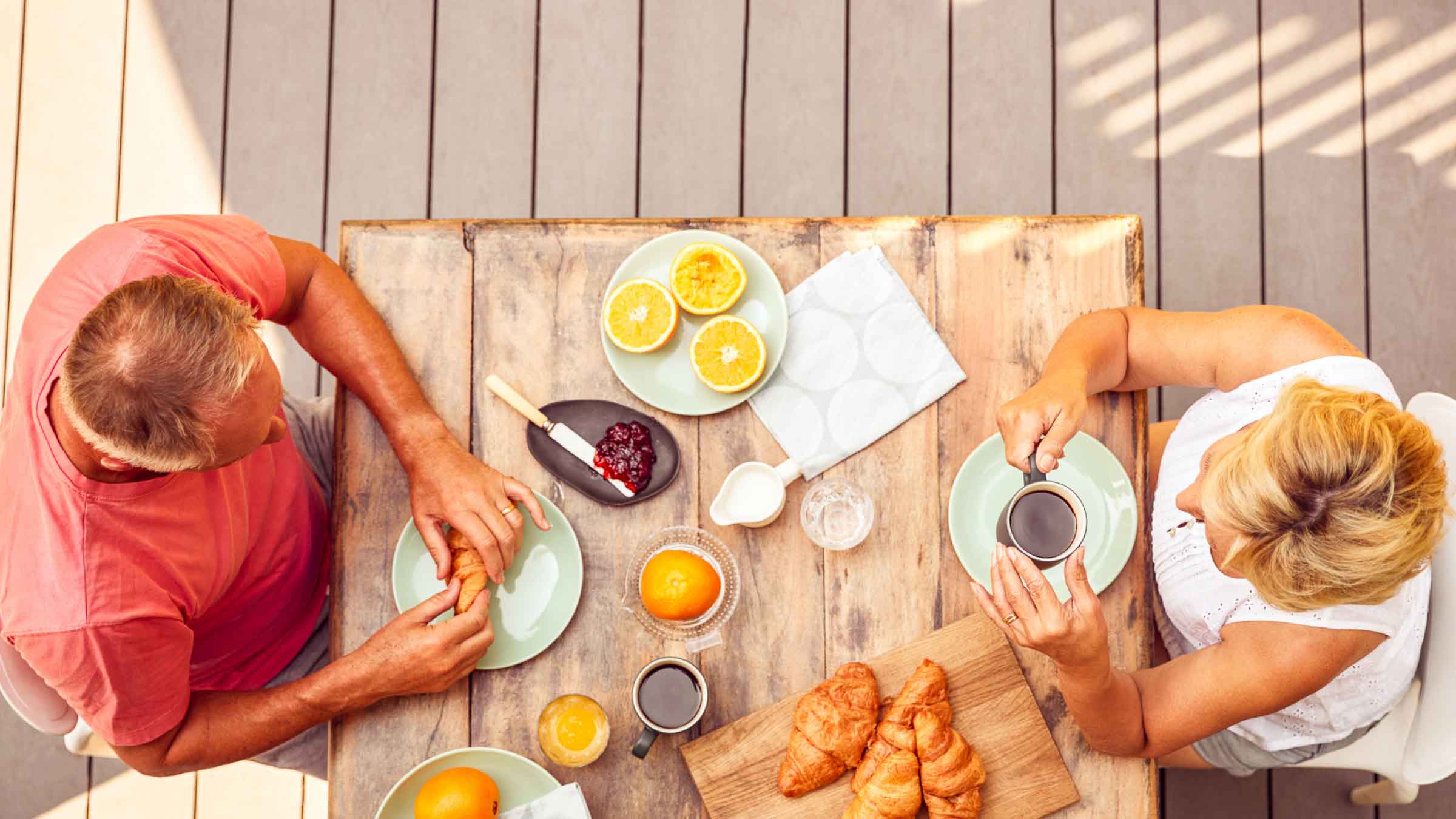 Ariel view of a couple sitting outside on their wooden decking, feasting on freshly cut oranges and croissants.