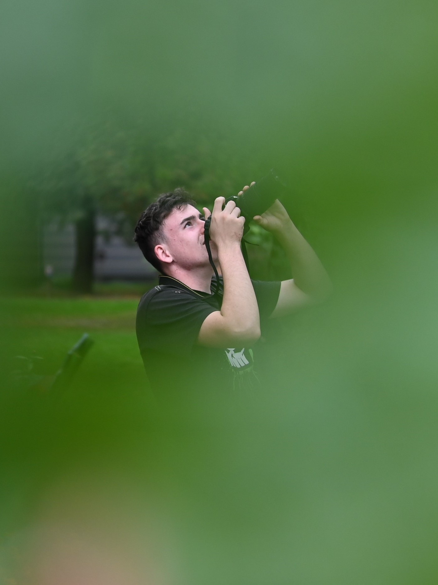 Warsztaty fotograficzne | Kadr złapany przez dziurkę  liściu, dokoła widać tylko rozmazane, zielone tło, a przez otwór z rozmytymi brzegami widać profil chłopca przykładającego aparat do policzka i patrzącego w górę..JPG