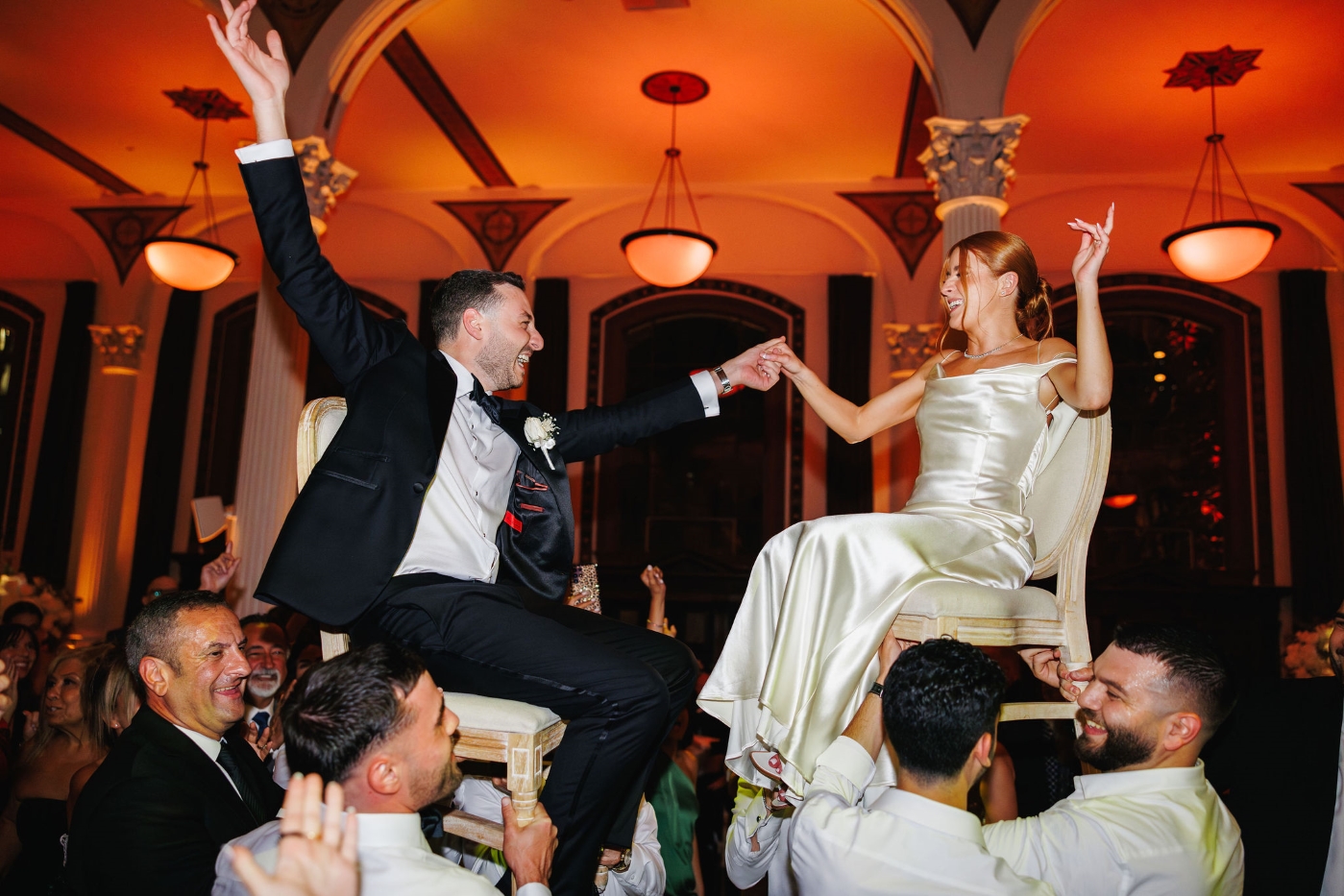 a bride and groom being lifted up on chairs by their wedding guests, everyone is smiling