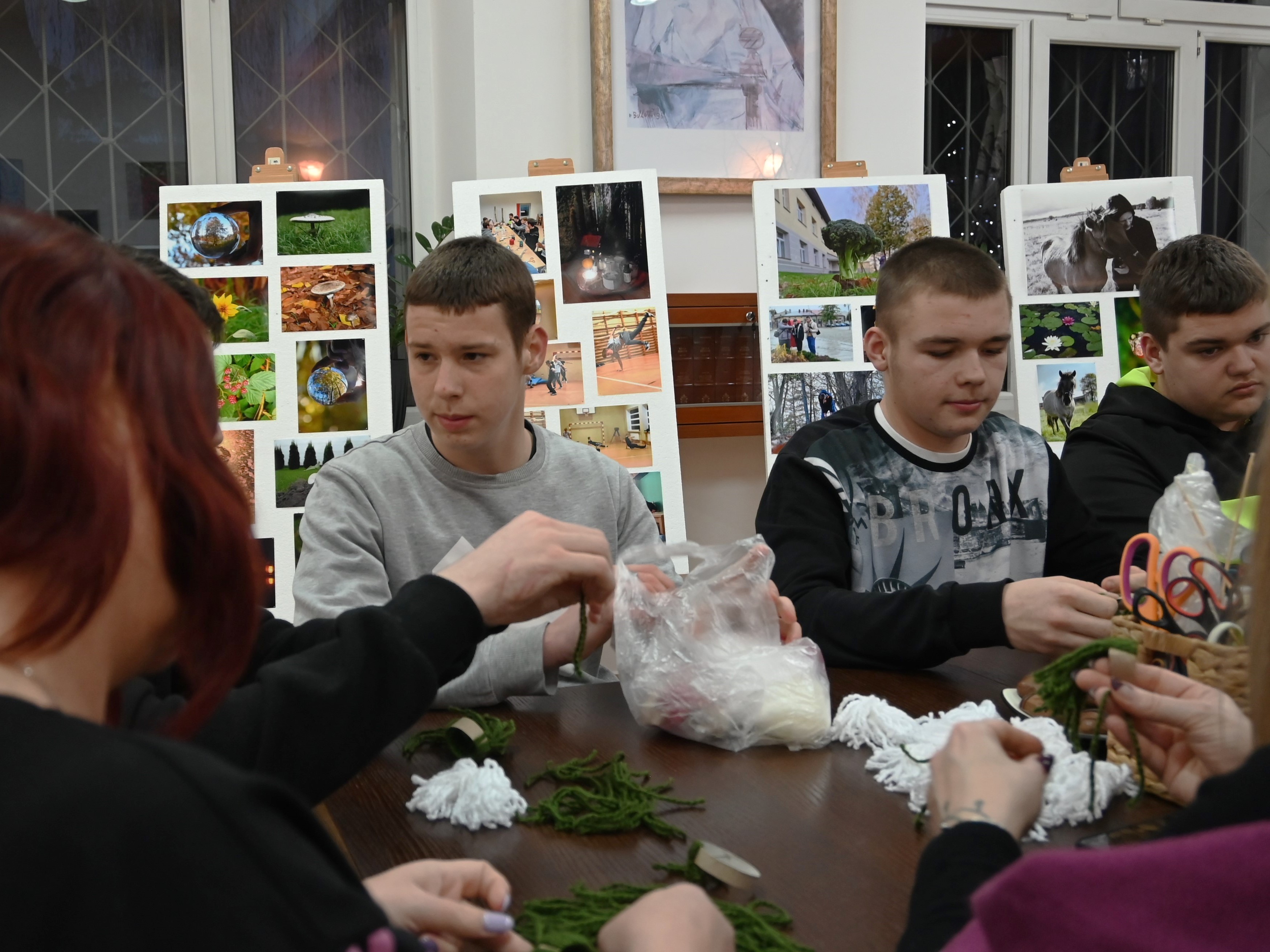 Zakończenie projektu Fajna Ferajna | Trzech chłopców siedzących przy stole, z boku widoczne włosy i bark kobiety. Na blacie stołu leżą zielone i białe sznurki, foliowa reklamówka, koszyk z nożyczkami.JPG