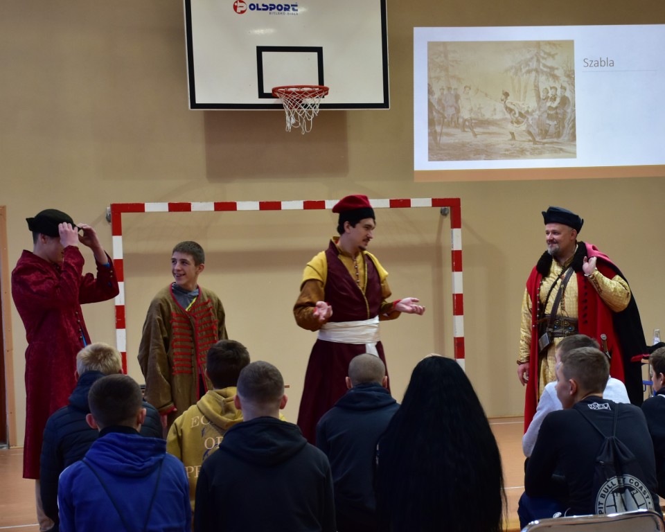 Lekcja historii | Trzech mężczyzn w kolorowych strojach szlacheckich, między nimi wychowanek przebrany w szlacheckie ubranie. Przed nimi siedzą oglądający wychowankowie MOW. W tle na ścianie wyświetlana prezentacja multimedialna..JPG