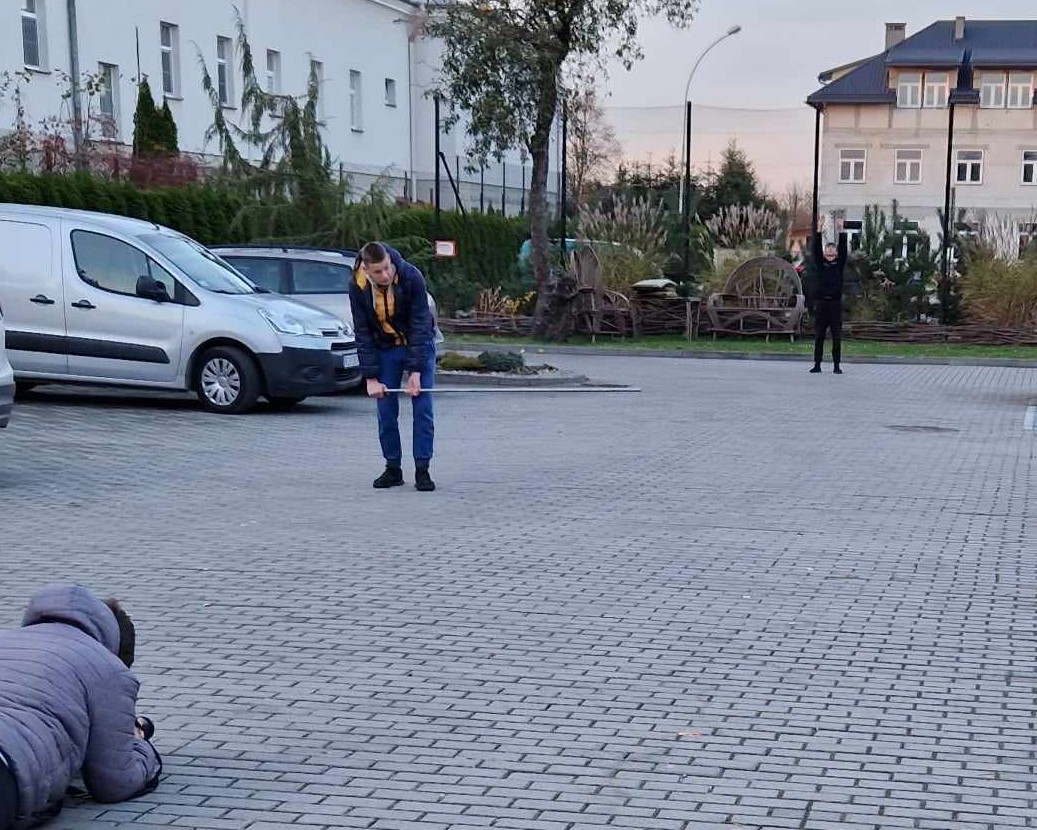 Cykl zajęć z programu „Fajna ferajna”  | Plac wyłożony kostką, na nim dwóch chłopców pozuje do zdjęć w perspektywie stojąc daleko od siebie. Na pierwszym planie w rogu leży inny chłopiec robiąc pozującym zdjęcie. W tle auta, budynki, drzewo.jpg