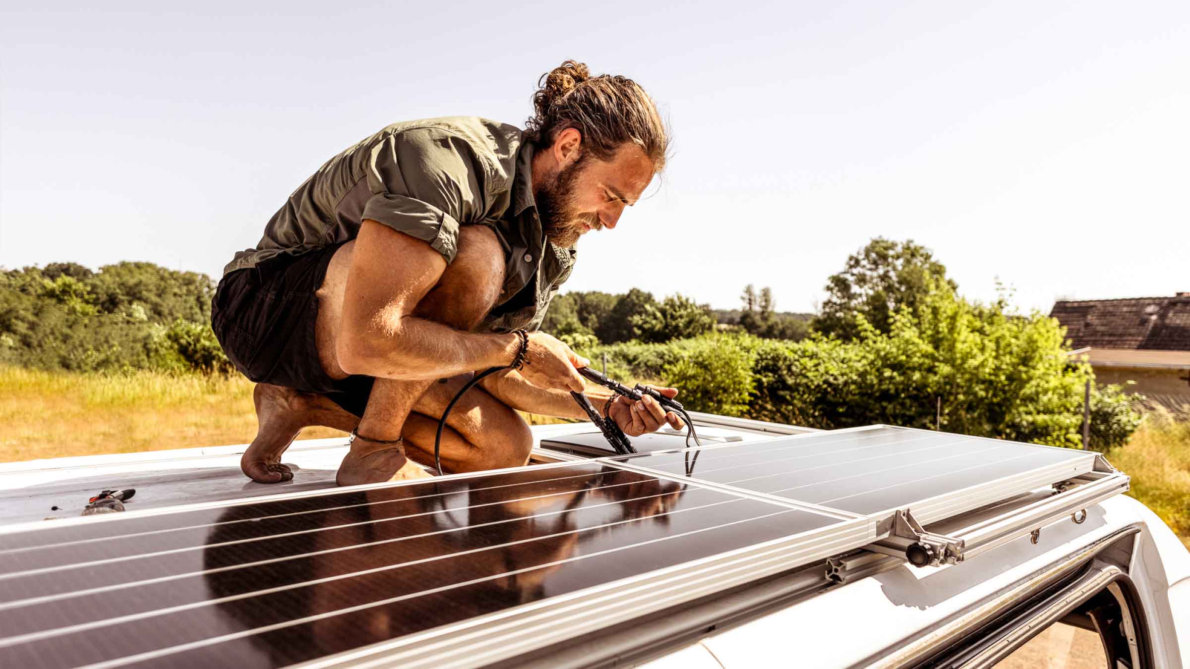 Young man crouched down on one knee, inspecting solar panel wires on-top of his van. Surrounded by green and gold bushland.