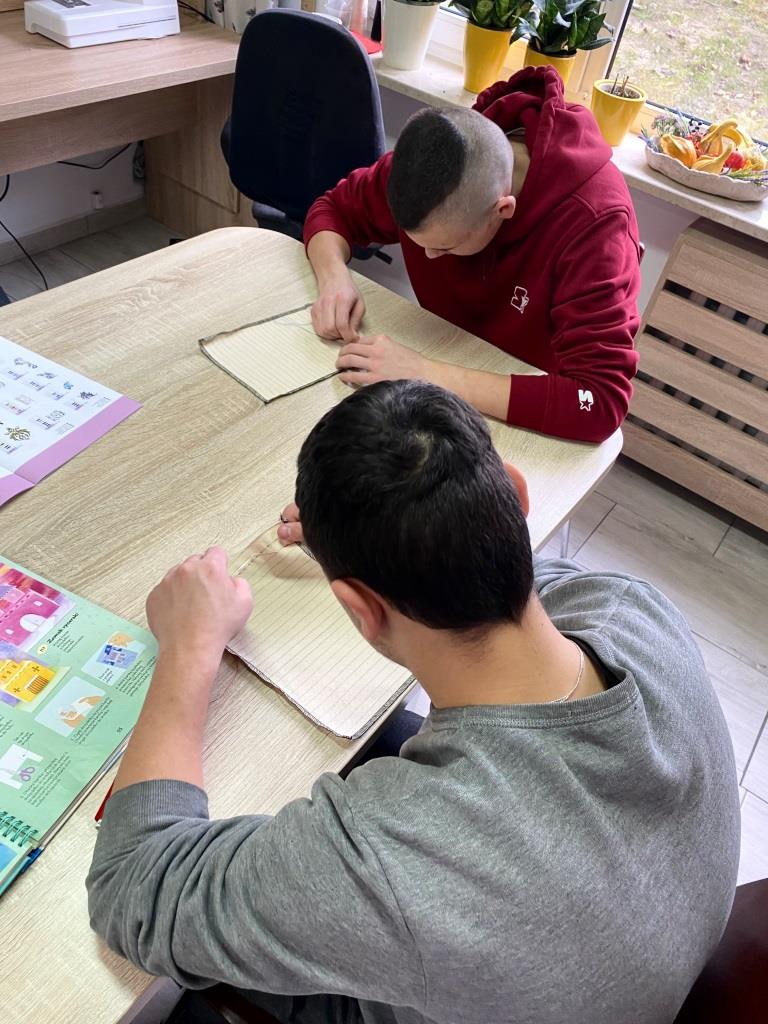 Wizyta w Warsztatach Terapii Zajęciowej w Oleszycach | Dwóch chłopców siedzi przy stole, przed nimi czyste kartki papieru. Zdjęcie z góry.jpeg