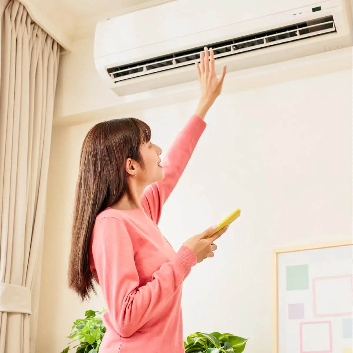 Woman in pink long sleeve top reaches up towards her air conditioner with remote in hand, to check if it's turned on.