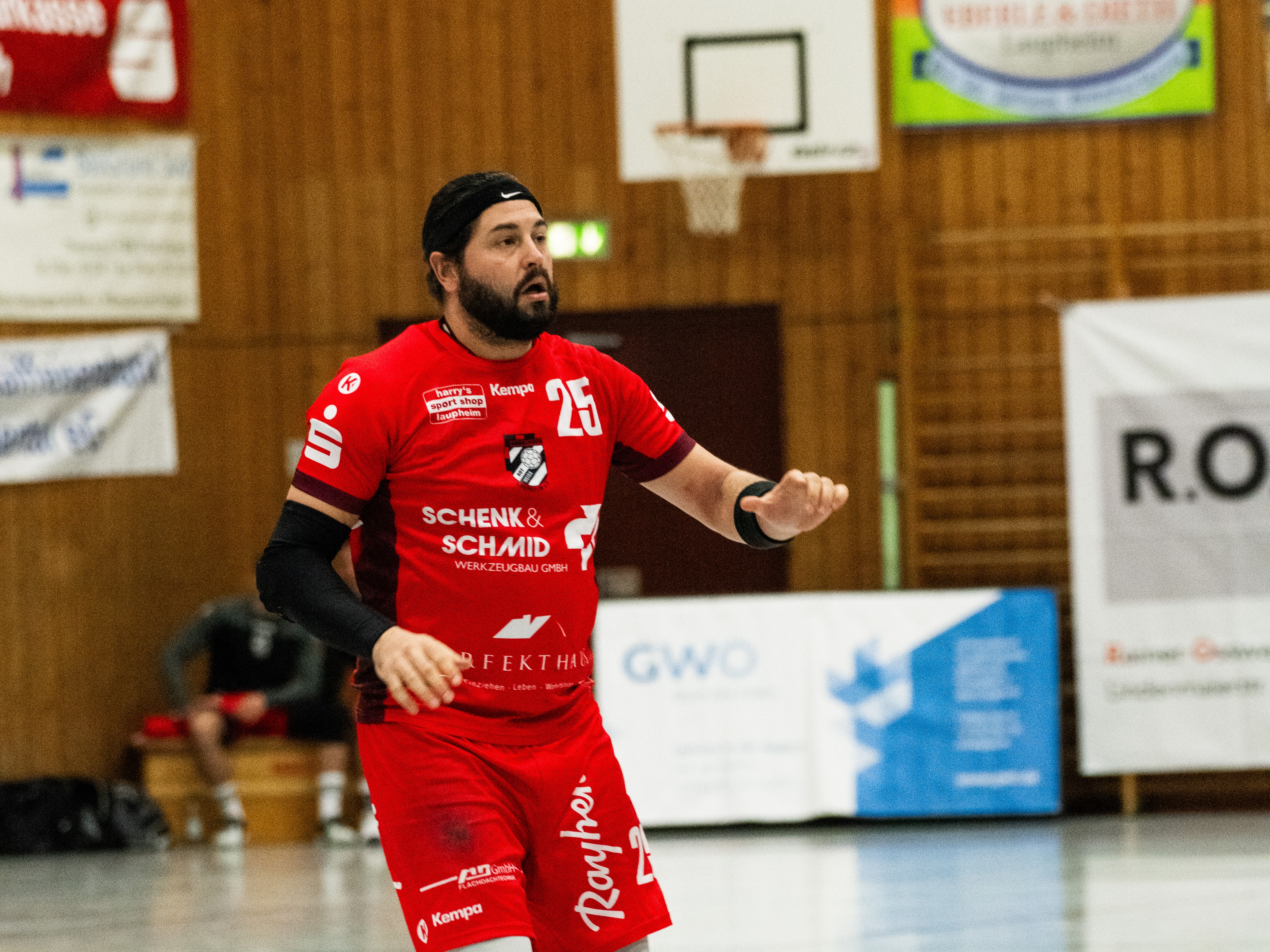 Spieler mit der Nummer 25 des HRW in rotem Trikot in der Halle. Im Hintergrund sind die Holzwand der Halle und einige Werbebanner zu sehen.