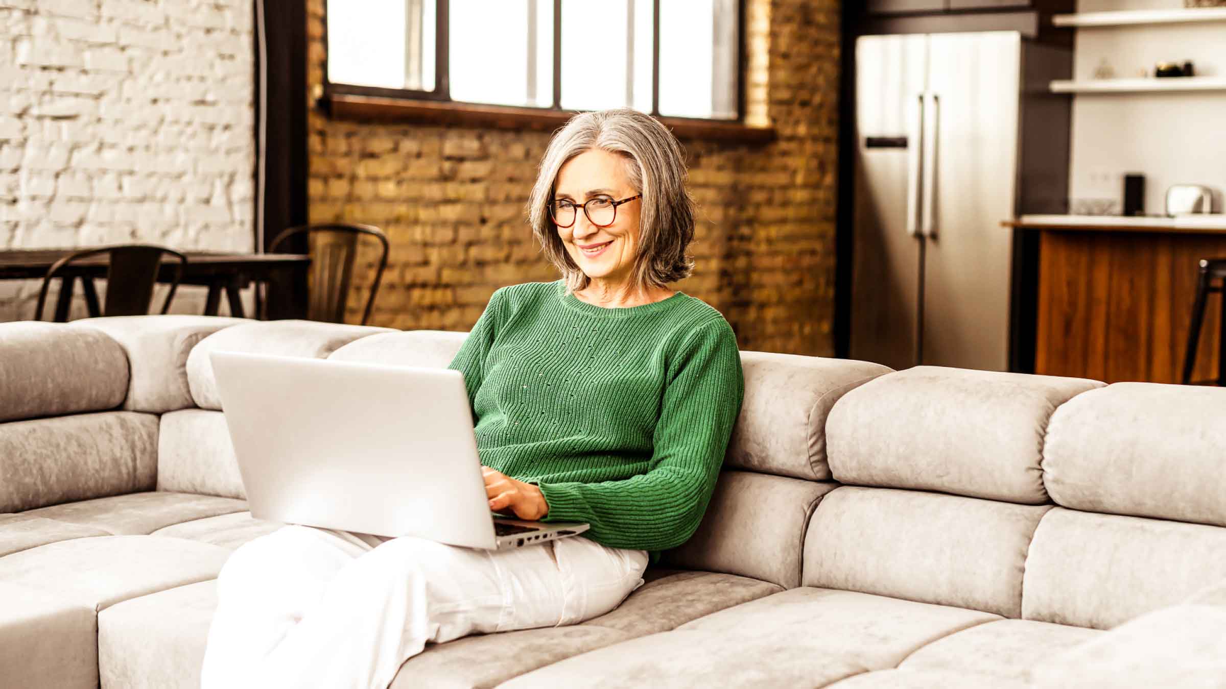 Smart woman with glasses and green jumper sits on sofa while looking at her laptop perched on her lap.