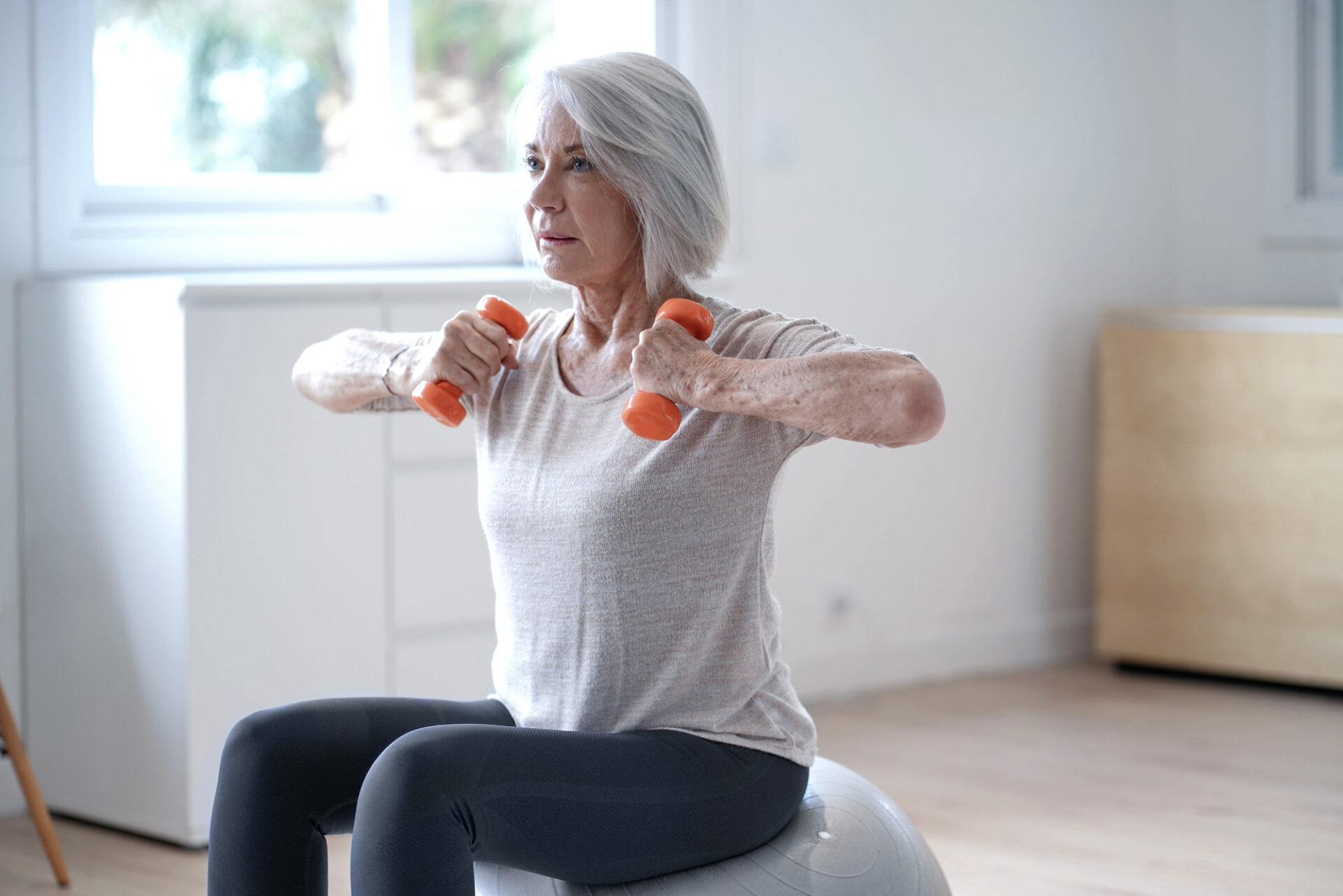 An older woman sitting on an exercise ball and lifting 2 dumbbells.