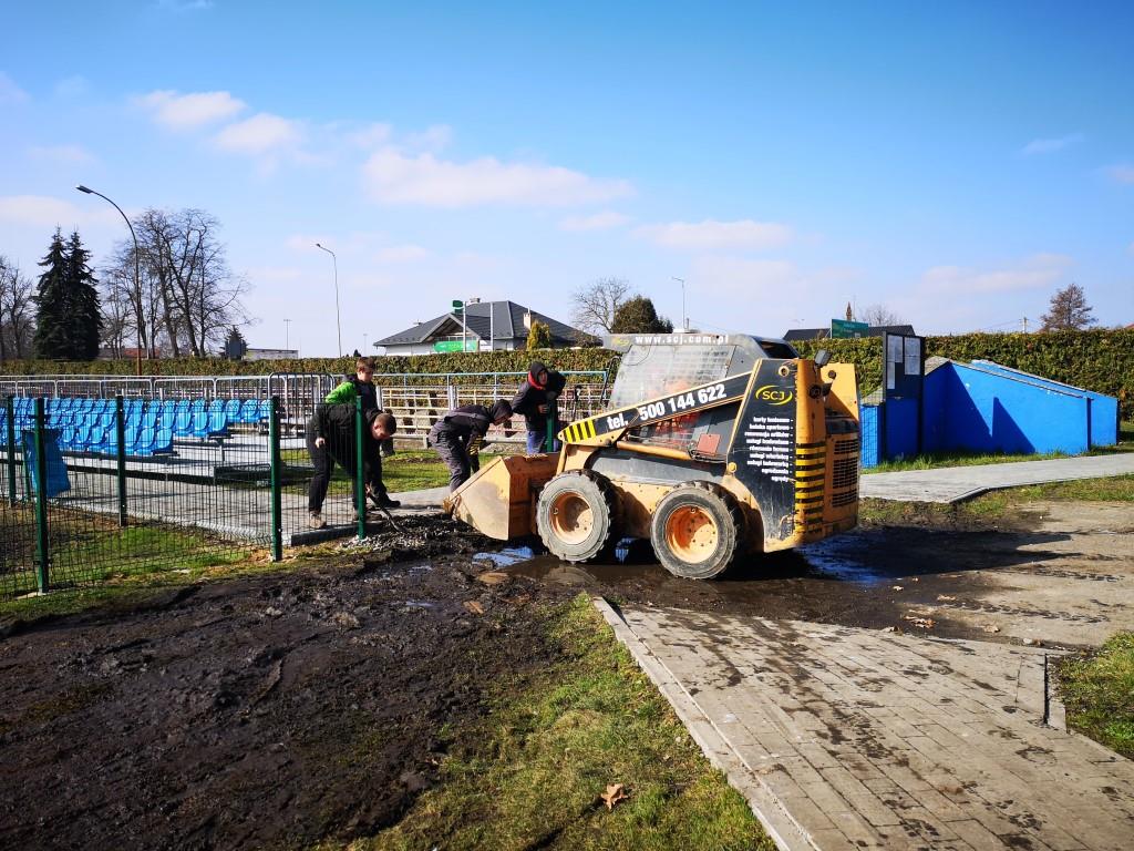 Porządki na stadionie | Na rozrytej ziemi stoi mała koparka z opuszczoną łyżką. Kilka osób ładuję na nią gruz.jpg