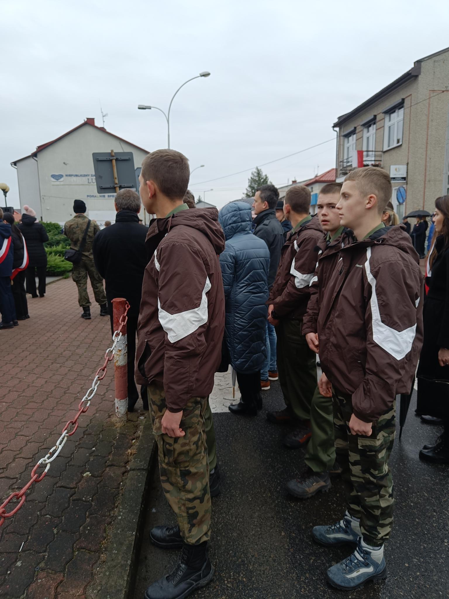 Narodowe Święto Niepodległości | Grupa chłoców w brązowych kurtkach stoi między innymi ludźmi podczas uroczystości pod pomnikiem. W tle zabudowania miasta..jpg