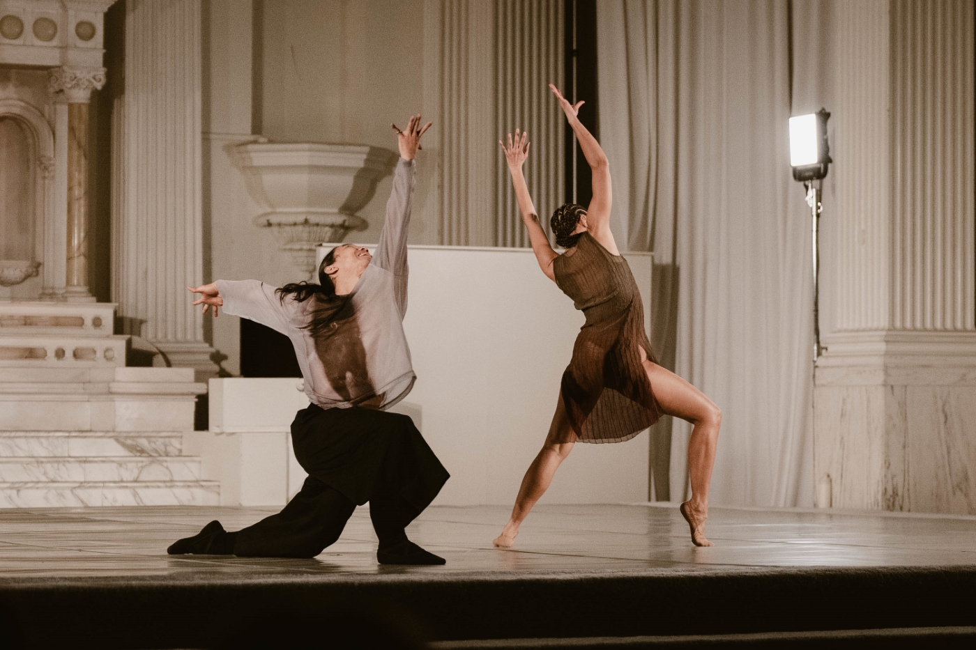 two dancers mid-performance with their arms outstretched toward the sky. one of the dancers is kneeling, at a dance performance at Vibiana in Downtown Los Angeles