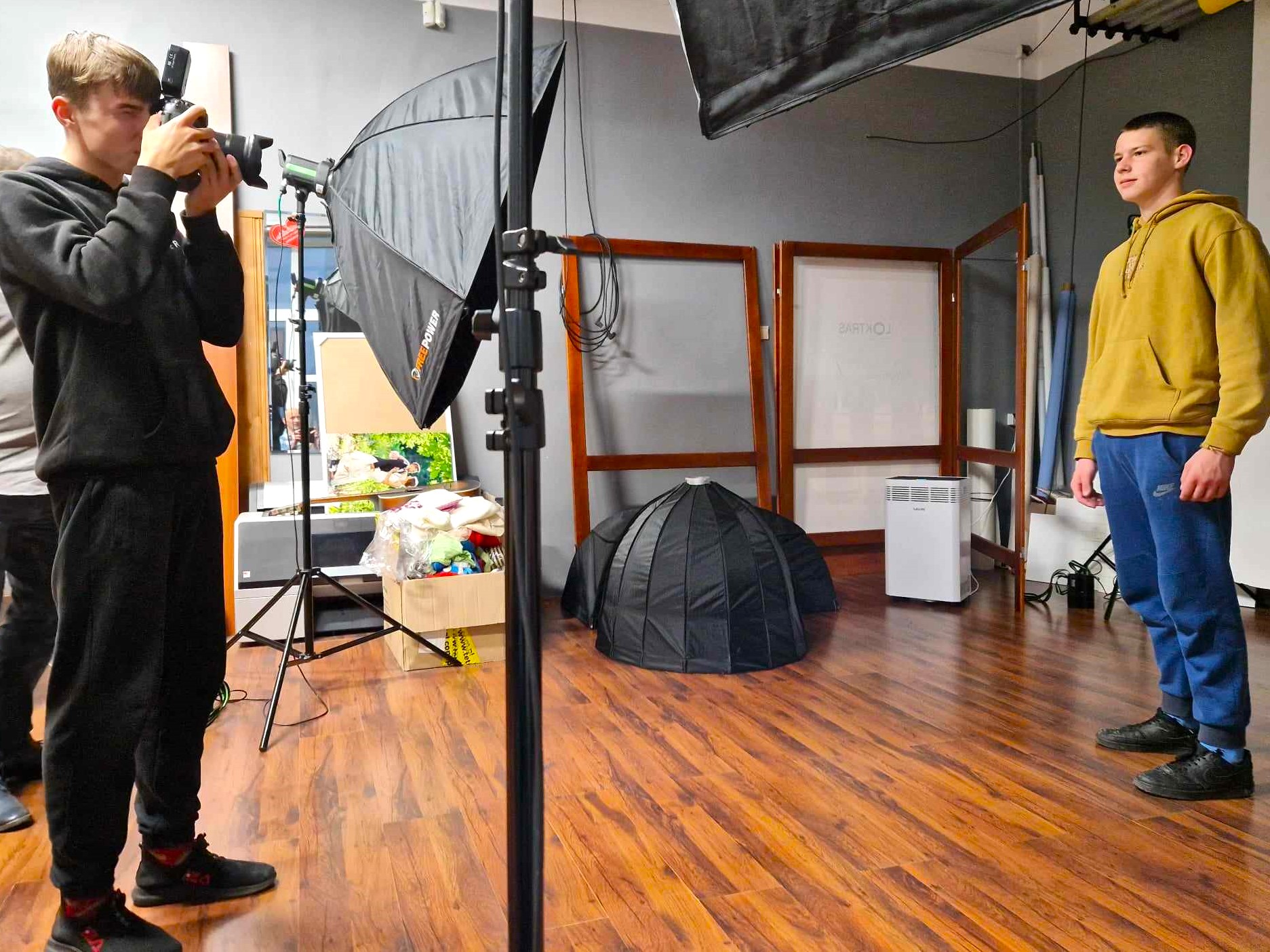 Zajęcia w studio fotograficznym | Chłipiec w czarych dresach stoi bokiem przykładając aparat do twarzy i robi zdjęcie koledze w żółtej bluzie i dżinsach. Wokól nich sprzęty i wyposażenie studia fotograficznego - lampy na statywach, rekwizyty..jpg