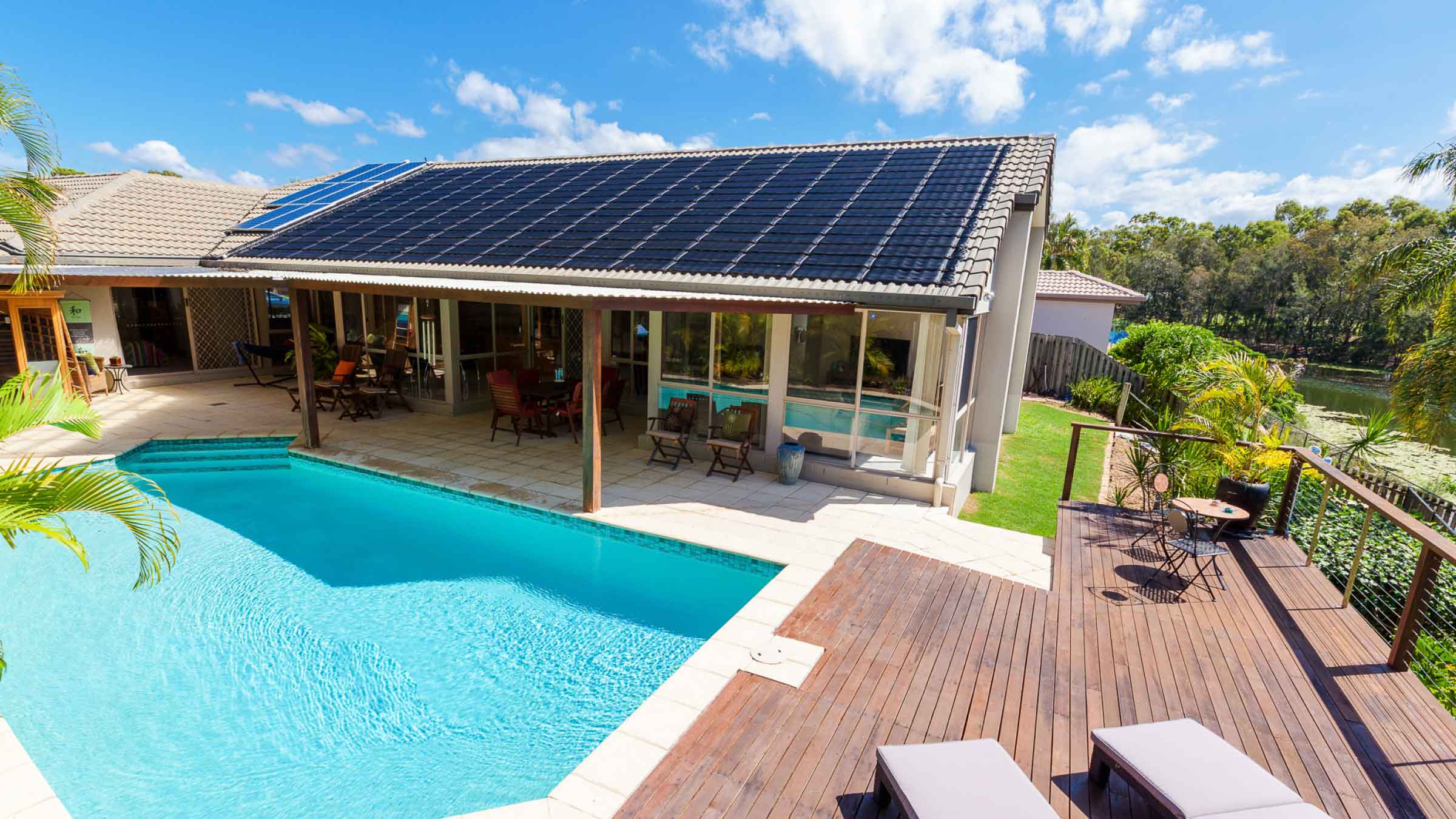 Solar tiles and panels on top of a modern bungalow, overlooking a pool with neat decking and manicured lawn.