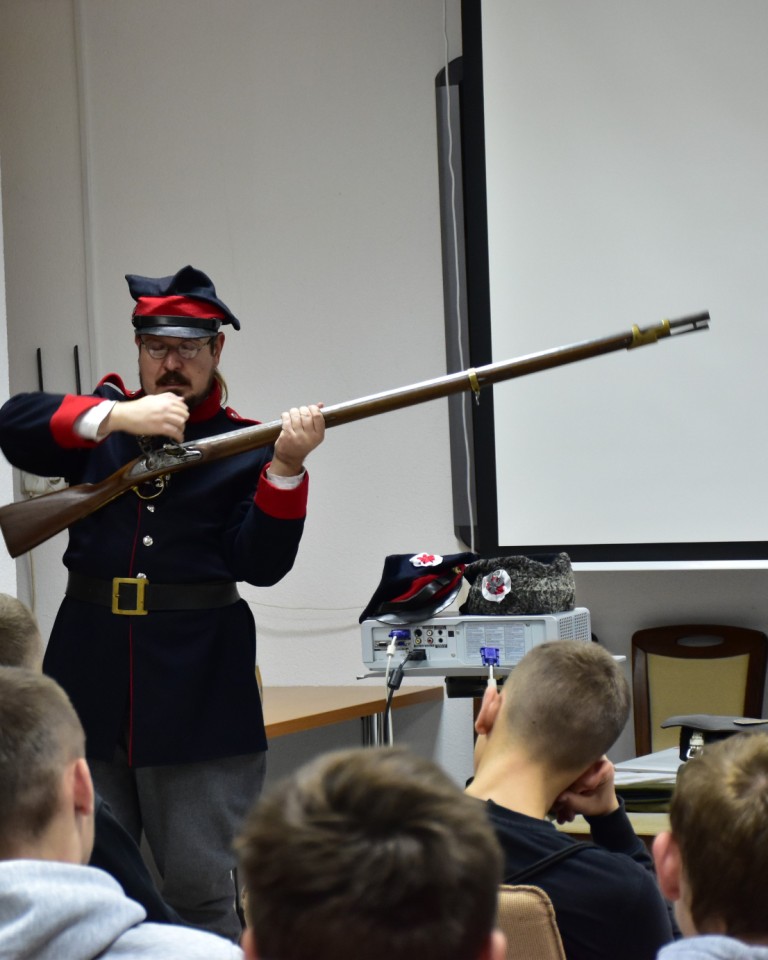 Lekcja historii | Mężczyzna w granatowym mundurze i czapce rogatywce oraz szarych spodniach prezentuje siiedzącym przed nim chłopcom broń palną - strzelbę z bardzo długą lufą..JPG