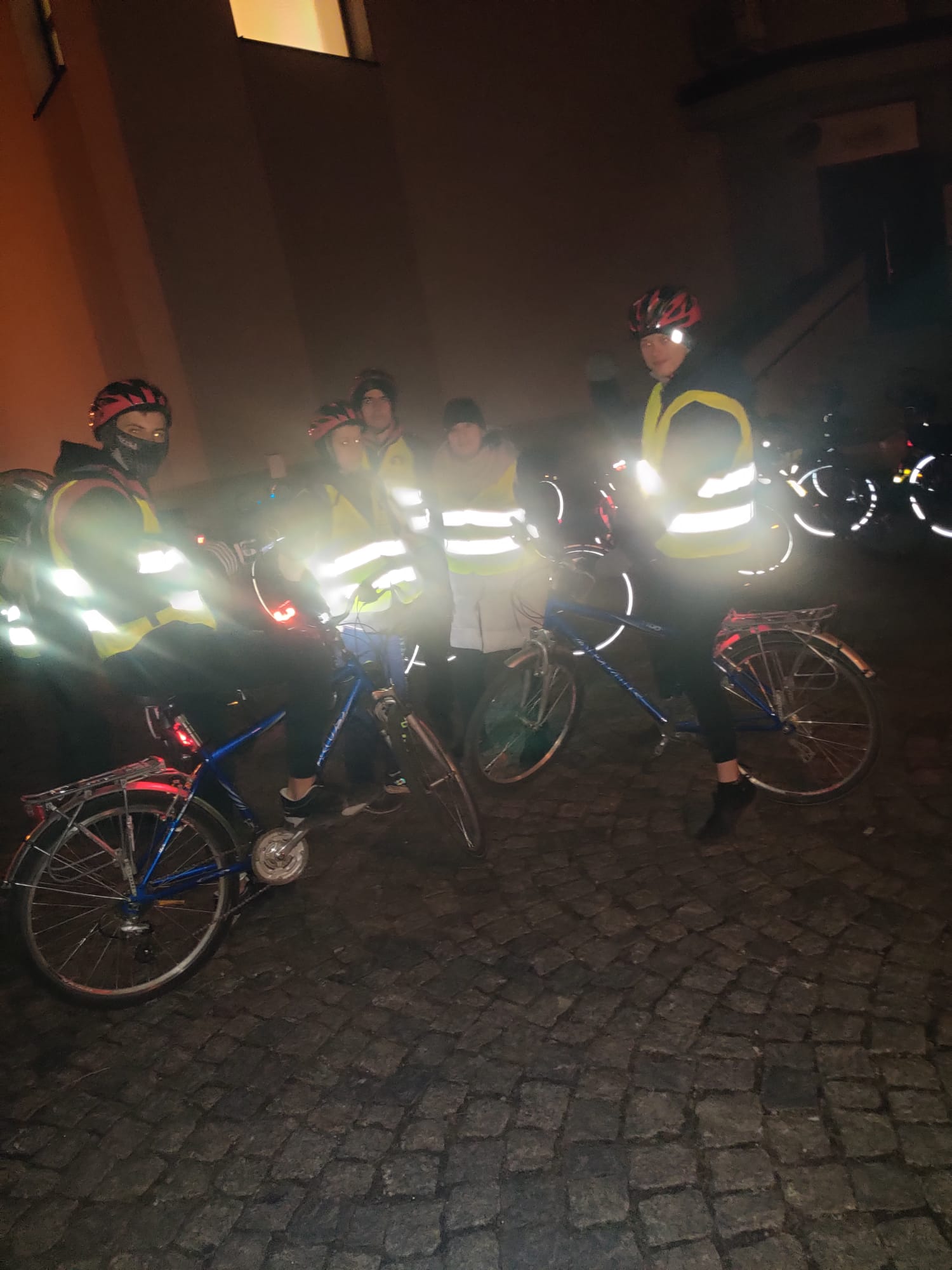 Rowerowa Droga Krzyżowa | Grupka kolarzy na rowerach stoi na brukowanym placu. W świetle lampy błyskowej bardzo mocno błyszczą odblaski na kamizelkach.jpg