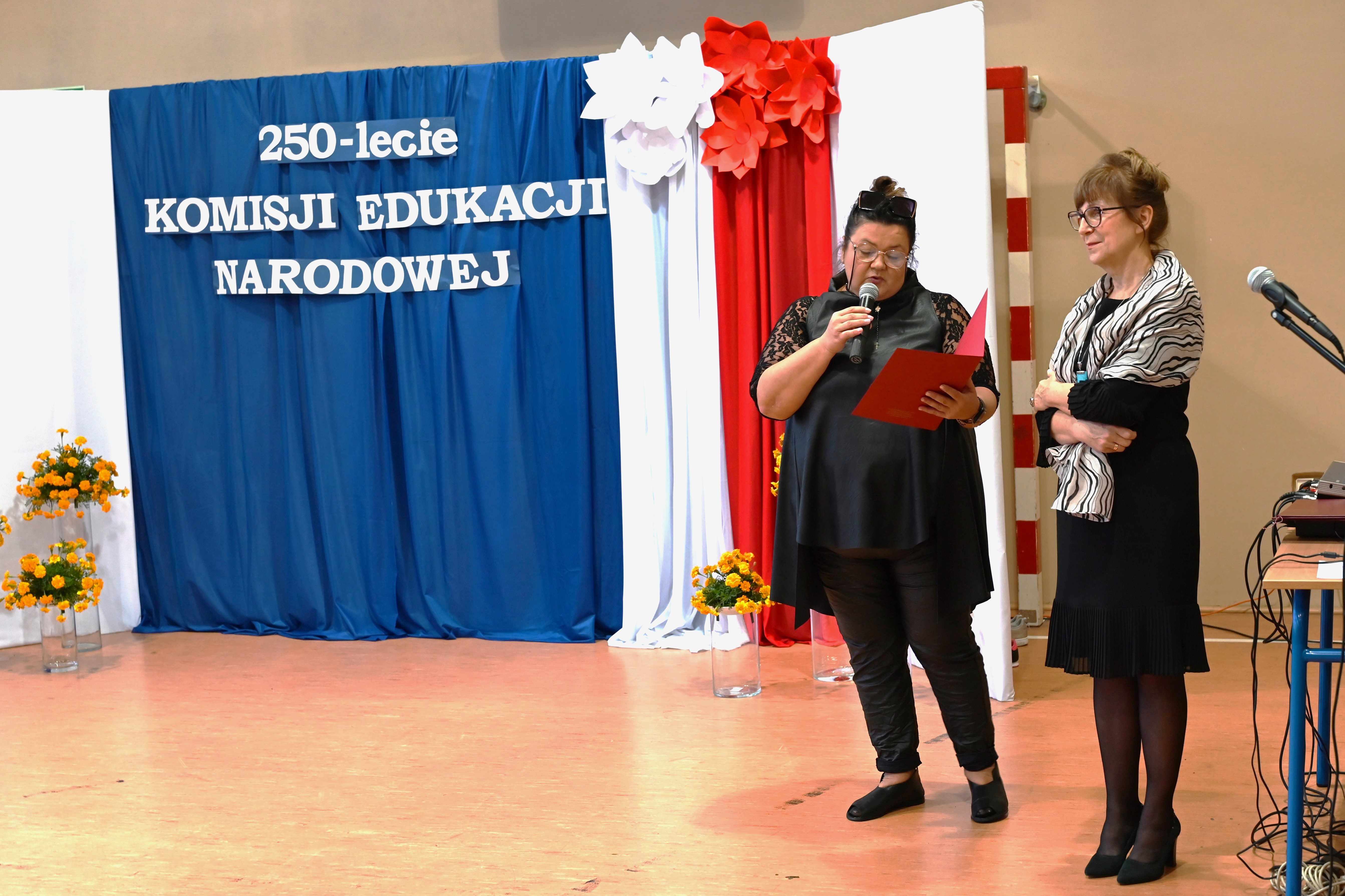 Dzień Edukacji Narodowej | Wice-dyrektorki SOSW, ubrane na czarno, stoją na tle dekoracji. Jedna z pań trzyma czerwoną otwartą teczkę, w drugiej ręce mikrofon i czyta..JPG