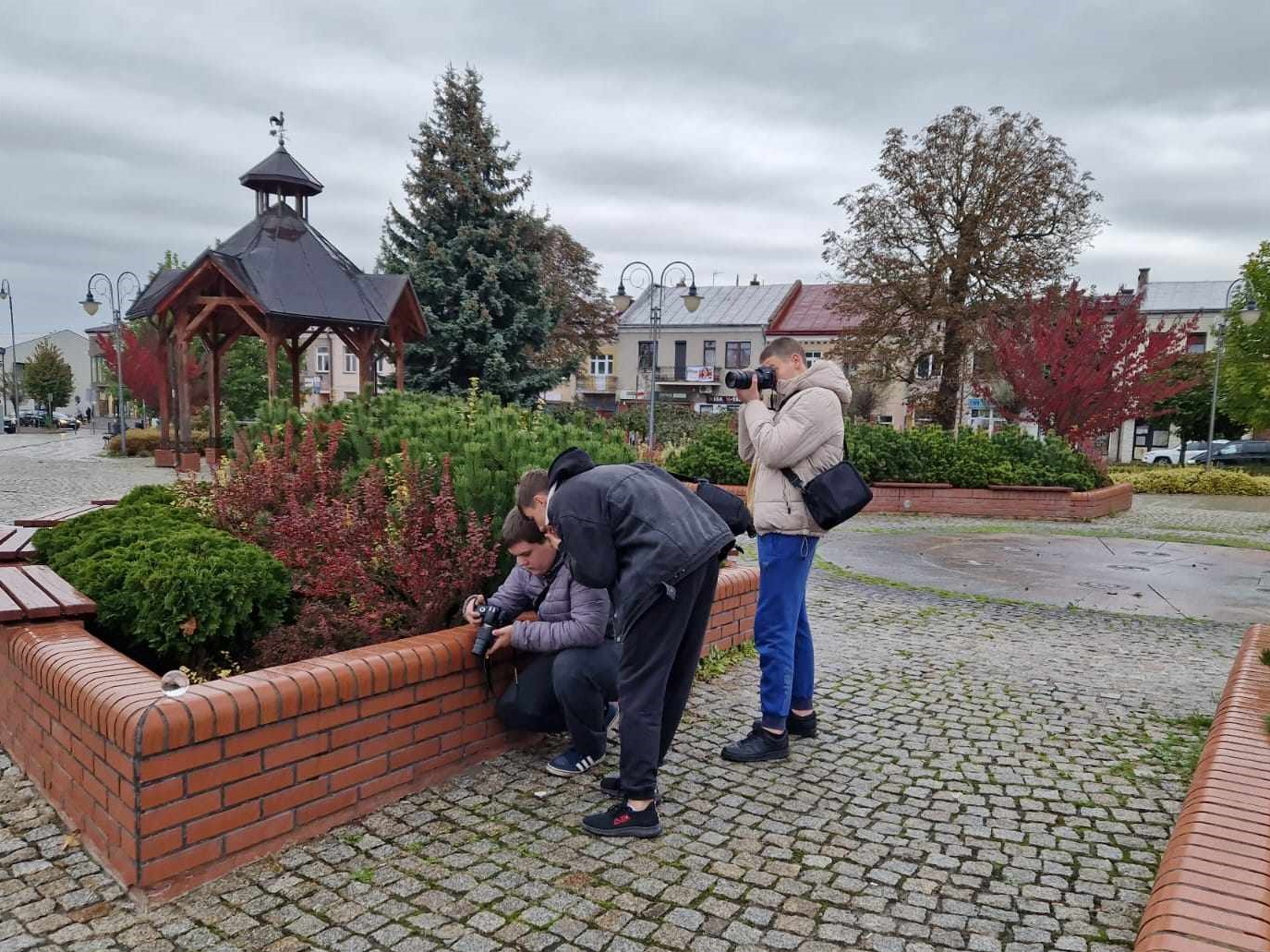 Fotografia jeszcze raz | Trzej chłopcy na Rynku w trakcie fotografowania. Obok nich ceglany murek ze szklaną kulą i jesienne krzewy. W tle zadaszenie miejskiej studni, za nią drzewa i kamieniczki..jpg