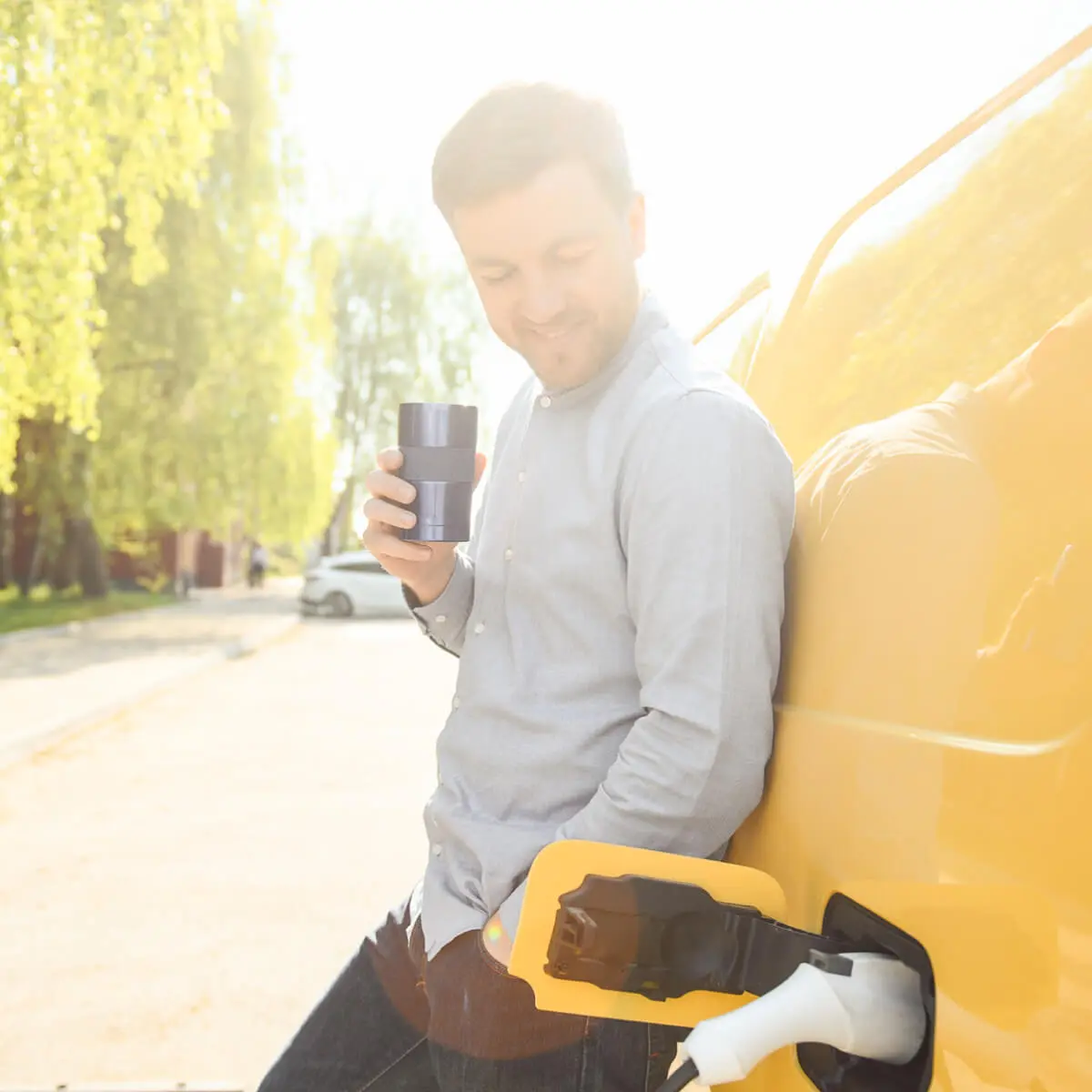 Young man, leans against yellow transit van drinking coffee, while waiting for it to electrically charge.