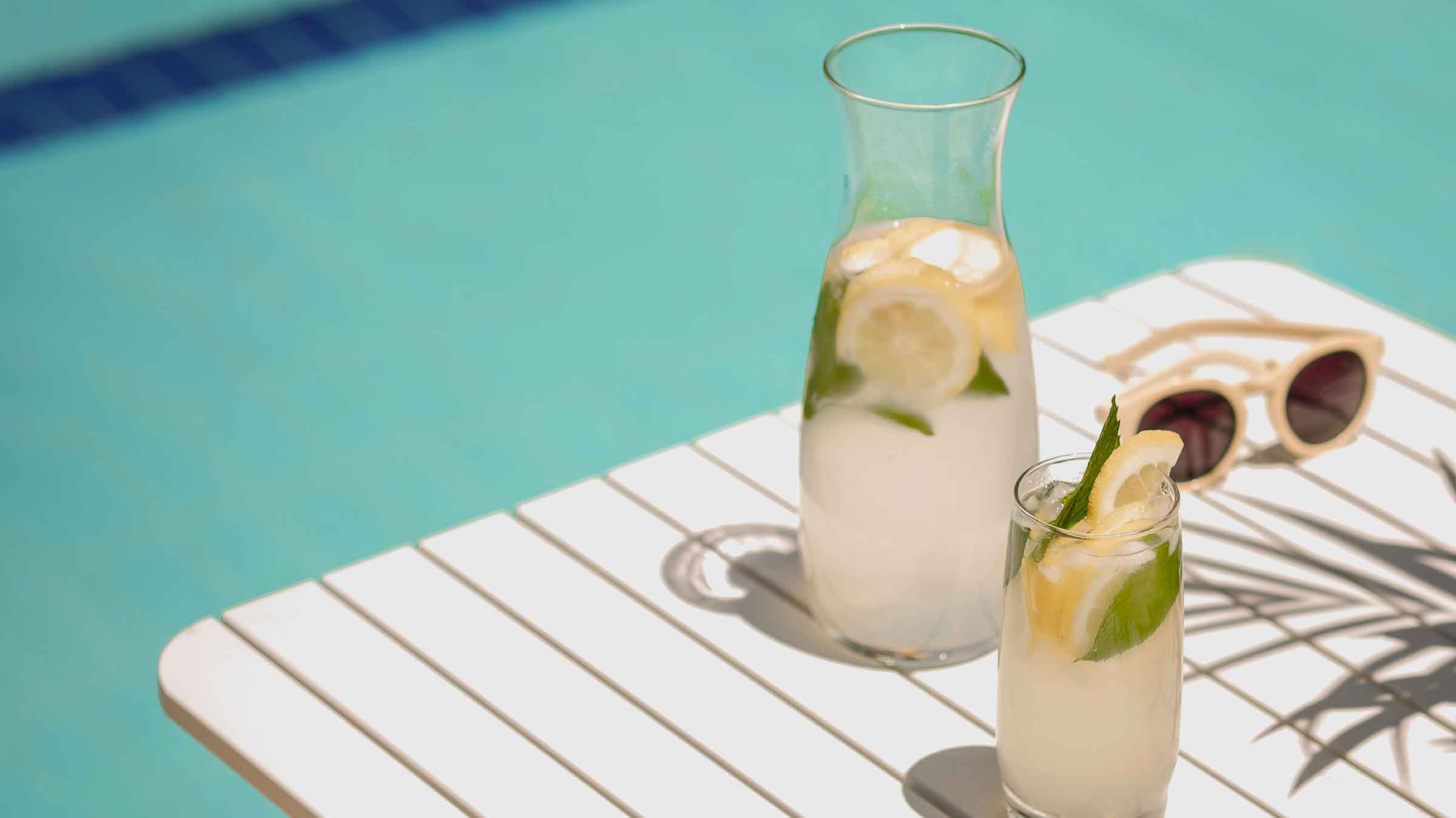 Jug and glass of lemonade garnished with slices of lemon and sprigs of mint, sit on top of a table beside a pool.