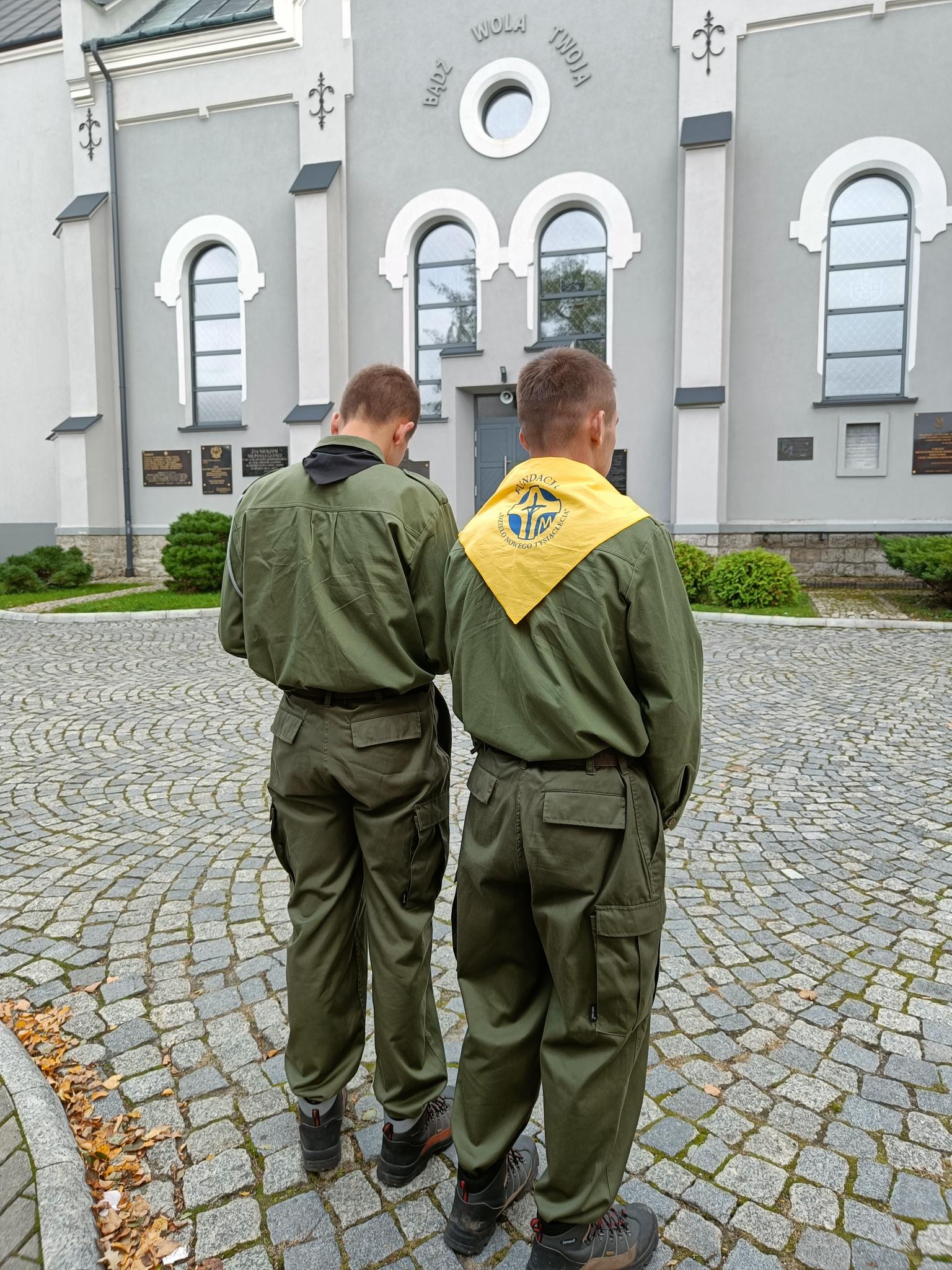 Dzień Papieski | Dwóch chłopców w harcerskich mundurach stoi tyłem na brukowanym placu kościoła, w tle fragment szarej bryły kościoła z półokrągłymi oknami..jpg