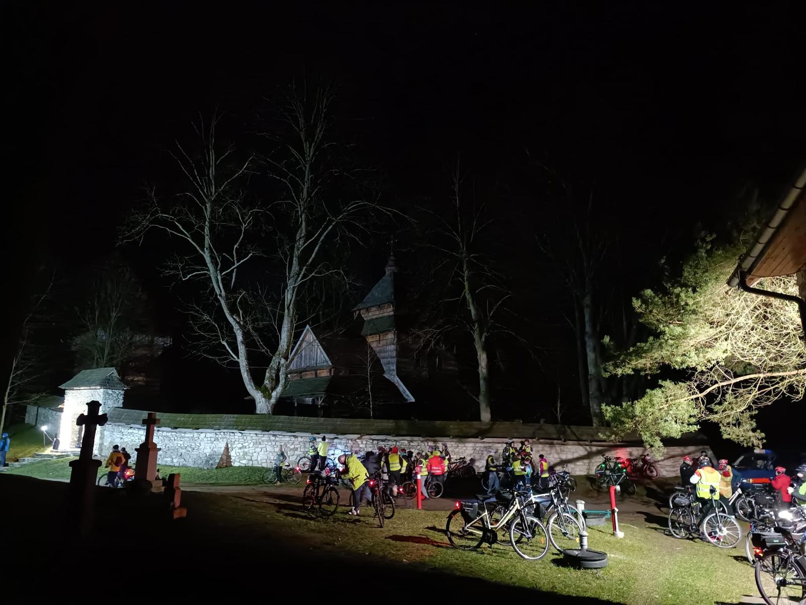 Rowerowa Droga Krzyżowa | Duża grupa kolarzy stoi przed murem z kamienia. Za murem widoczny fragment cerkwi i pnie ogromnych drzew. Zdjęcie nocne.jpg