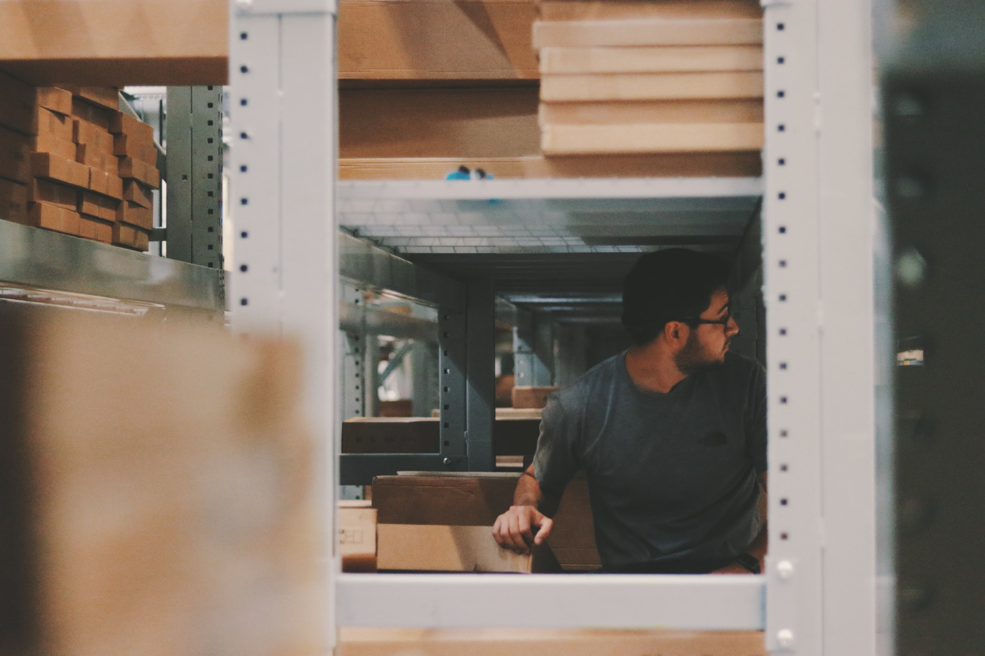 Blick durch Regale eines Lagerhauses auf einen jungen Mann mit Brille, der nach rechts schaut. Das E-Commerce Fulfillment ist für Onlinehändler ein entscheidender Erfolgsfaktor. Wir zeigen dir, auf was du achten solltest!