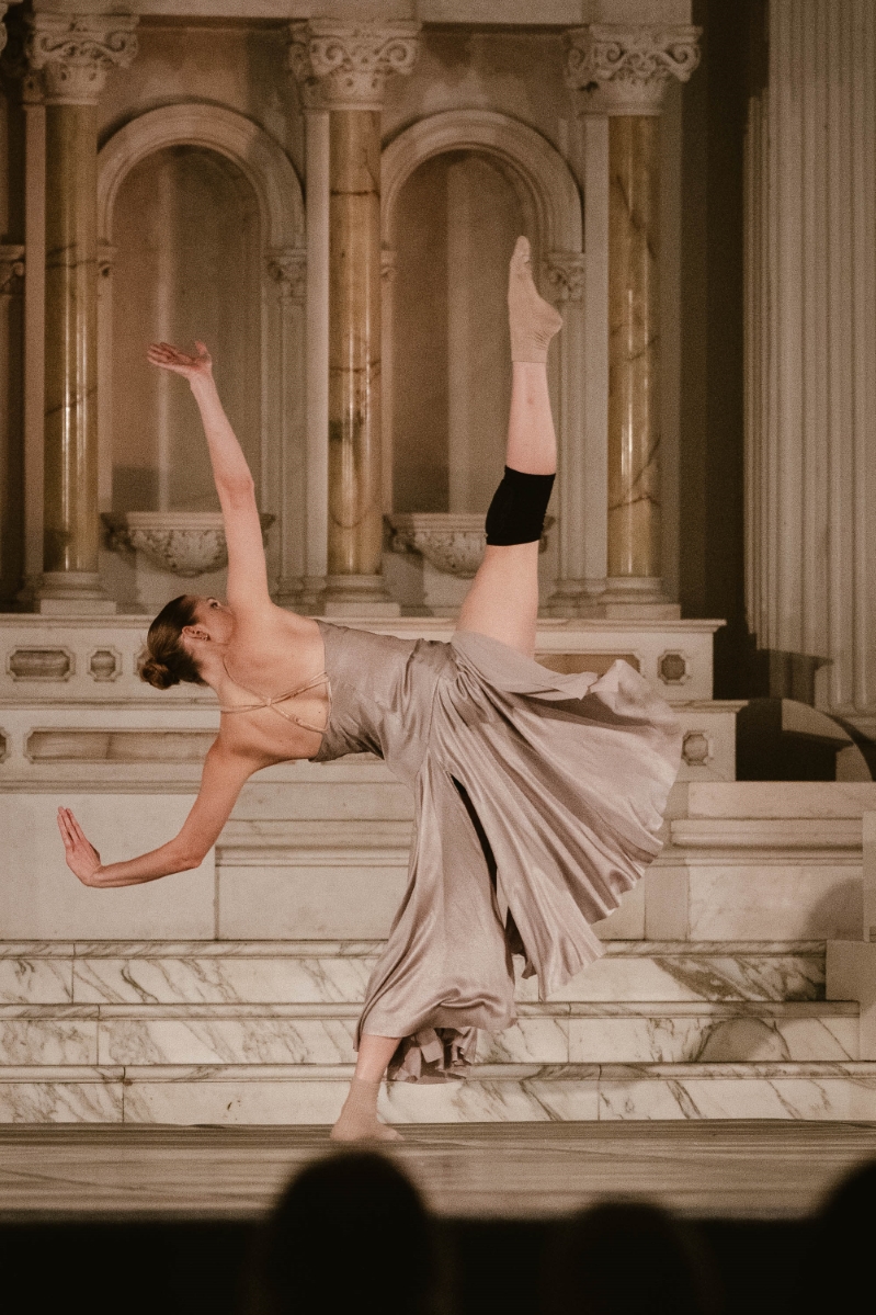 a female dancer in a neutral color dress points her toe toward the ceiling as she leans back, arms outstretched to keep her balance. at a dance gala performance at Vibiana in Downtown Los Angeles