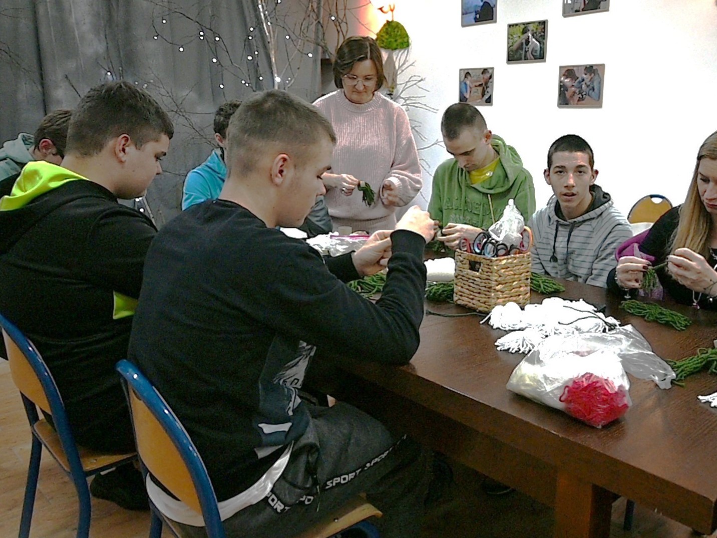 Zakończenie projektu Fajna Ferajna | Dokoła stołu siedzi młodzież, na blacie leżą kolorowe sznurki do plecionek. U szczytu stołu stoi wychowawczyni objaśniając splot.JPG
