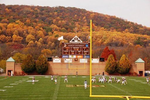 Students play a football game on a large field with a backdrop of fall foliage.