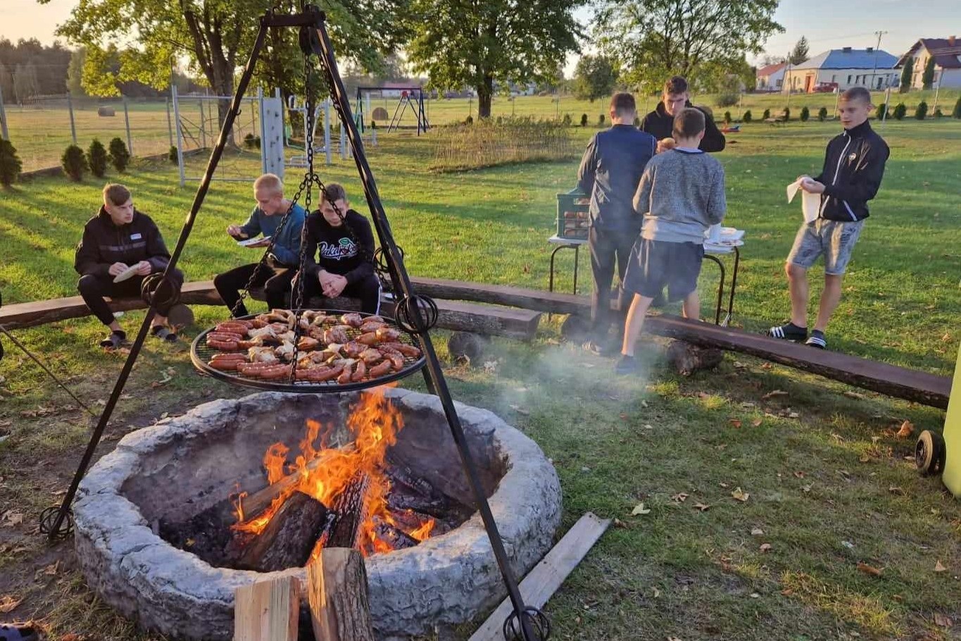 Obóz w Hucie Różanieckiej | Ognisko otoczone cembrowiną, nad nim ruszt z mięsem. Z tyłu ustwione w półkolu ławeczki, na których siedzą chłopcy. Część z nich stoi przy stoliku. Dokoła zieleń, w tle większe drzewa..jpg