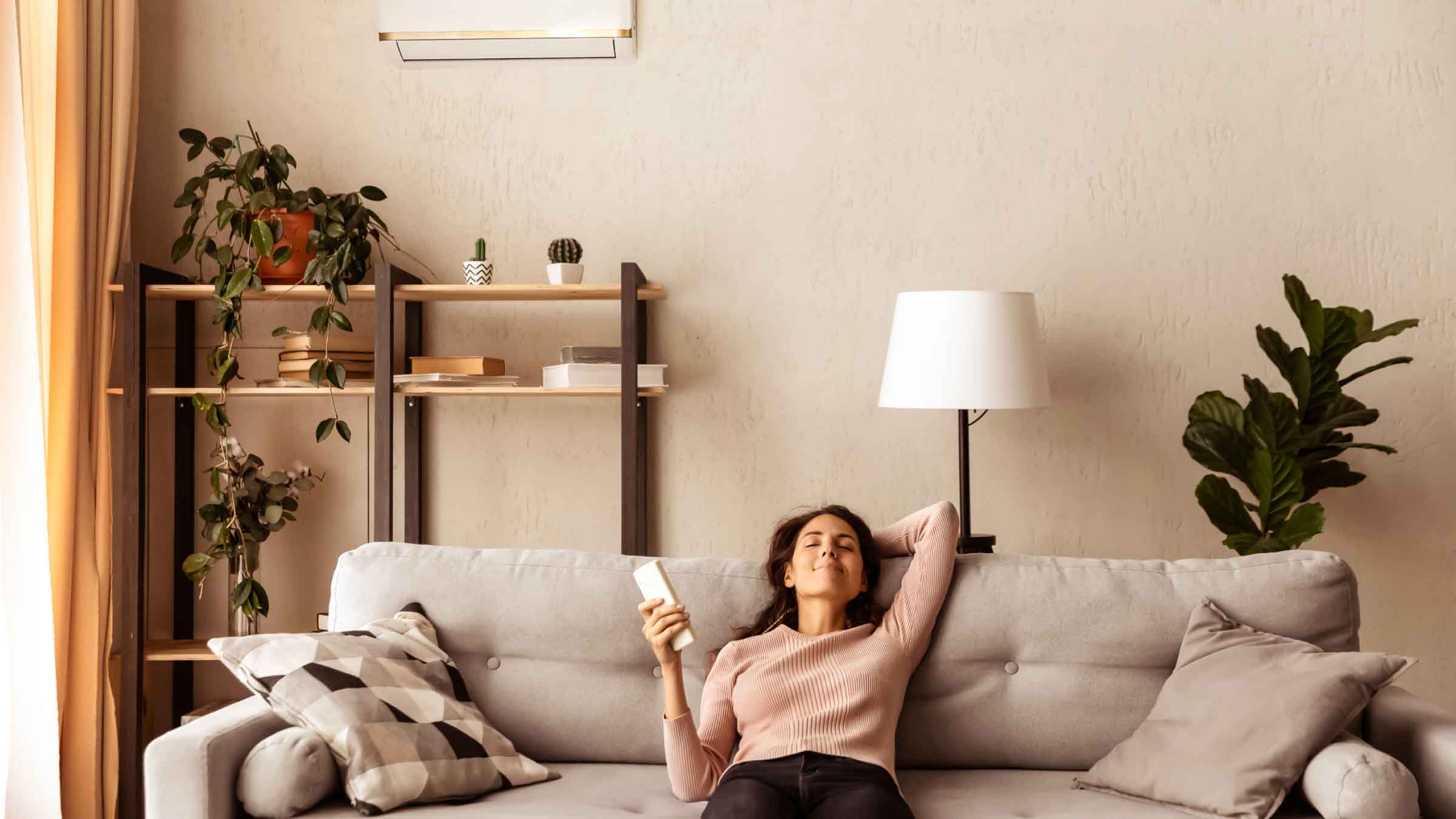 Woman relaxes back on sofa, one arm behind her head the other holds remote control pointed toward air conditioner.