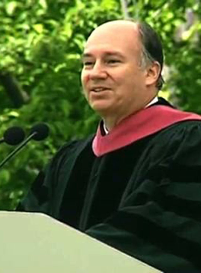 Karim Aga Khan IV wearing commencement attire, speaking at podium on outdoor stage, trees in background