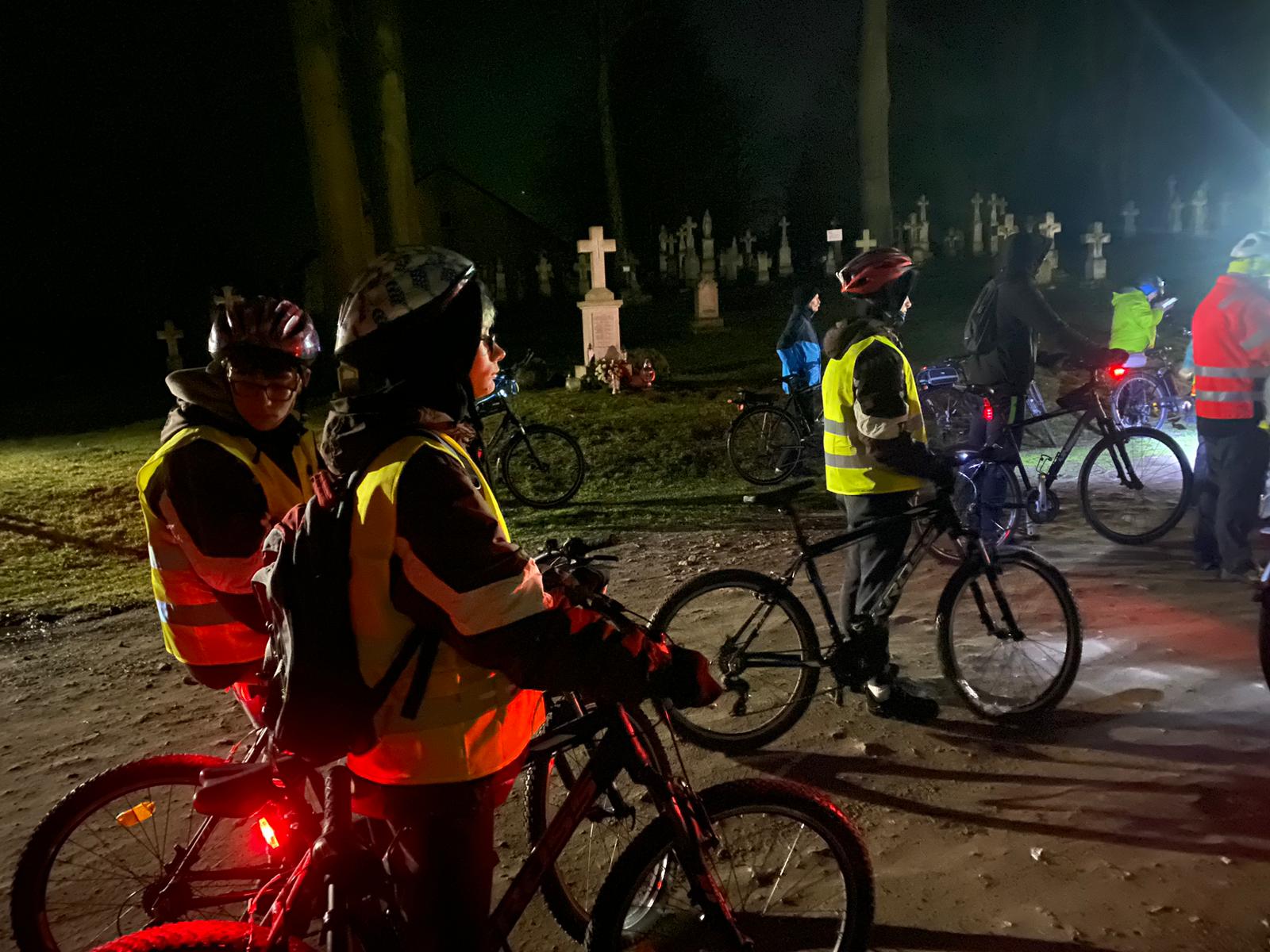 Rowerowa Droga Krzyżowa | Kilku wychowanków MOW stoi bokiem przy swoich rowerach. W tle widoczne kamienne krzyże nagrobków i pnie drzew. Zdjęcie nocne.jpg