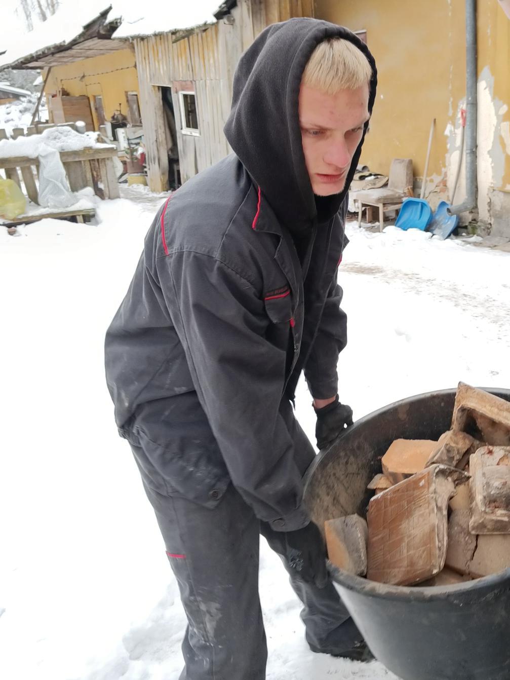 Wolontariat – pomoc w rozbiórce pieca | Chłopiec niesie wspólnie z kimś niewidocznym na zdjęciu  okrągły pojemnik wypełniony kaflami z rozbiórki. Dokoła śnieg, w tle ściana domu.jpg