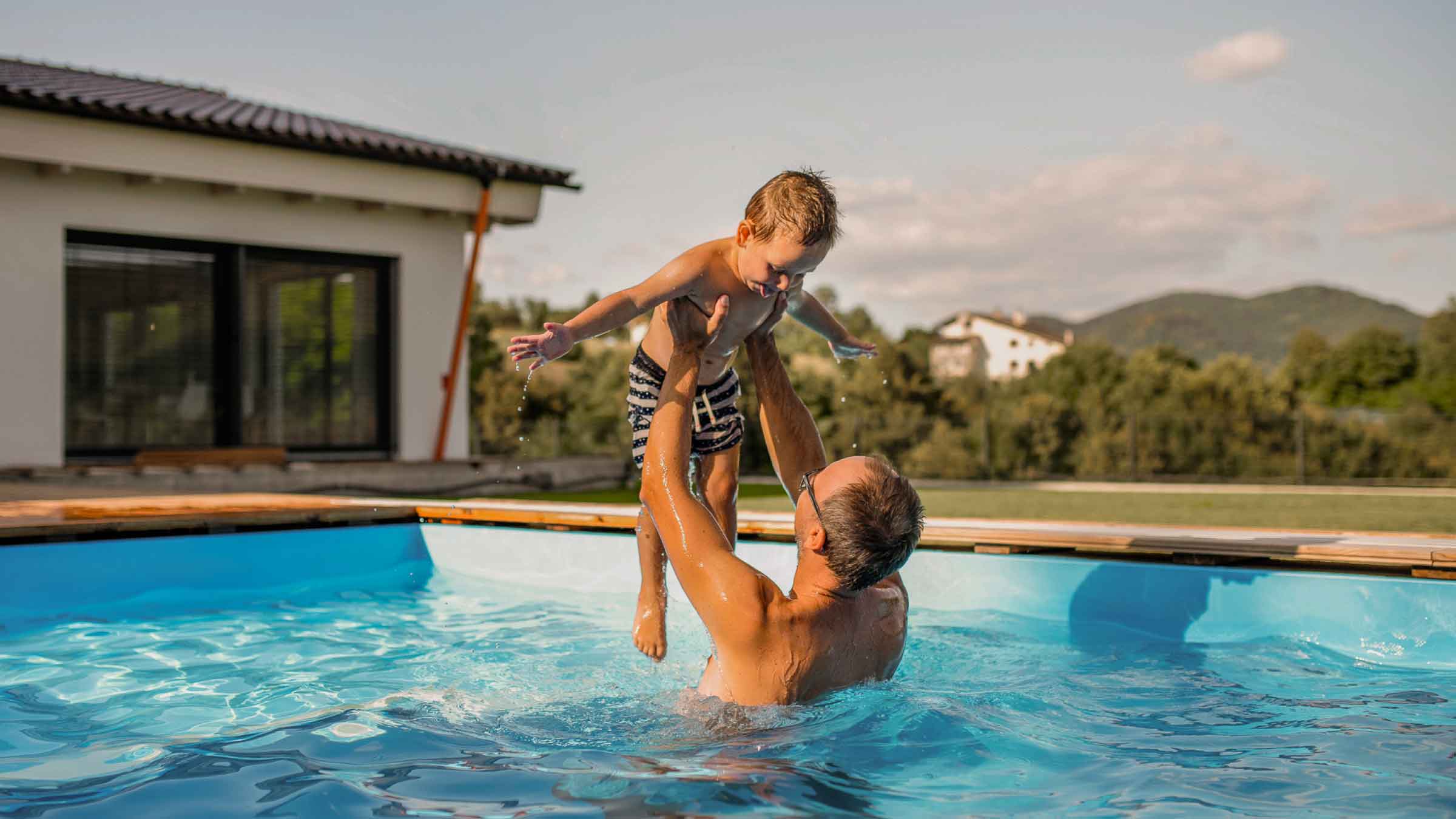 Parent standing in pool beside home, holds young child up above him, green hills surround them in the background.