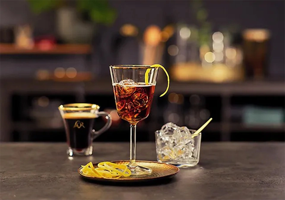 A chilled cocktail featuring a twist of lemon and a subtle backdrop of L'OR Espresso coffee graces the dark table.