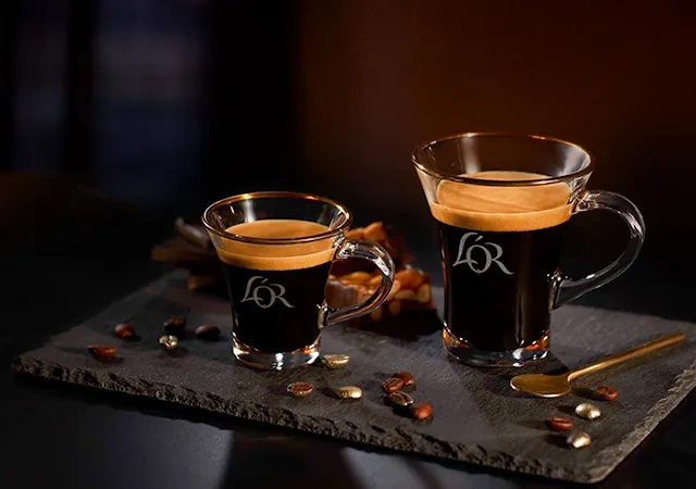 Two mugs filled with L'OR Espresso on a slate tray, with scattered coffee beans and a silver spoon for an elegant presentation.