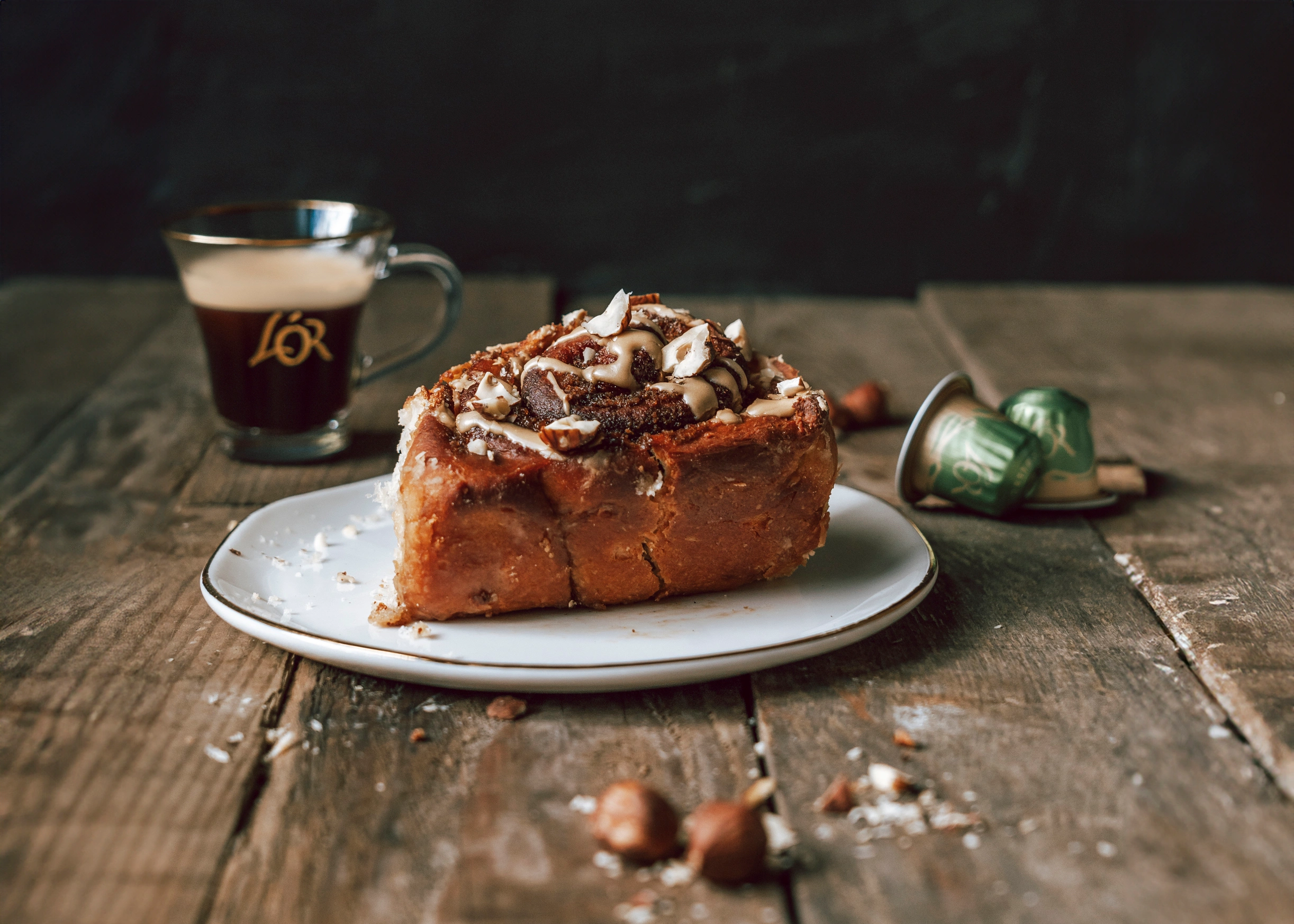 A cinnamon roll with nuts sits invitingly on a wooden table, accompanied by a steaming cup of rich L'OR Espresso or perhaps some other delightful variant from the L'OR range. The warm, aromatic coffee perfectly complements the sweetness and crunch of the pastry, creating an ideal pairing for any coffee enthusiast.