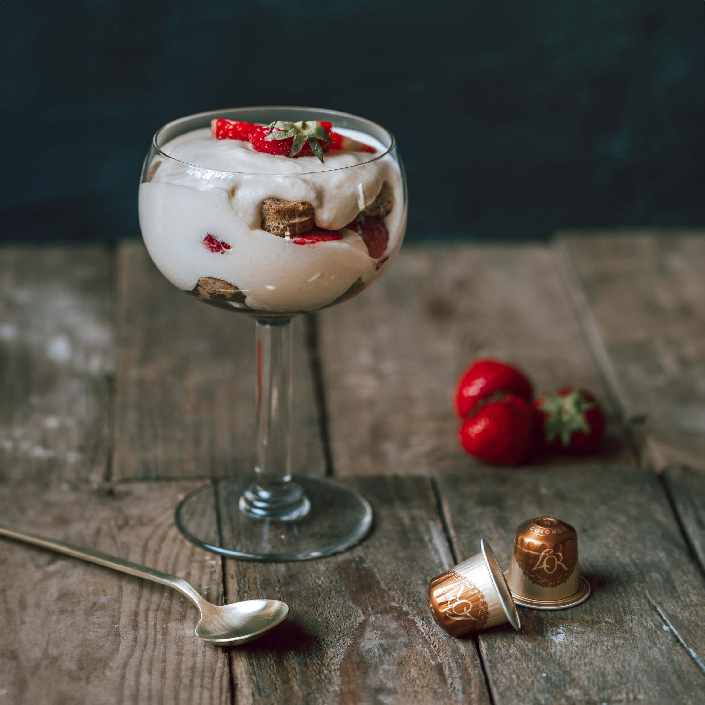 Tiramisu in a glass, elegantly layered with fresh strawberries and enriched by L'OR Espresso, sits invitingly on a rustic wooden surface. Adorned with coffee capsules, this dessert offers a harmonious blend of flavors and textures.