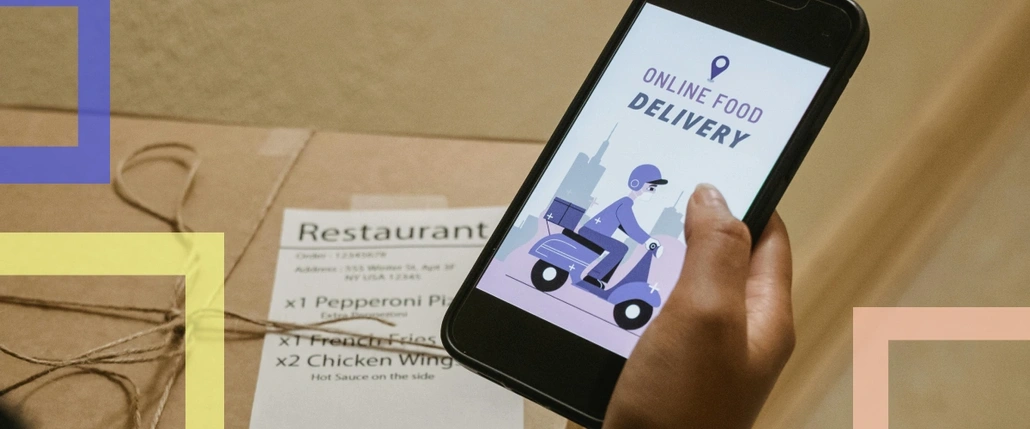 A box with food and a hand holding a smartphone with delivery app on the screen