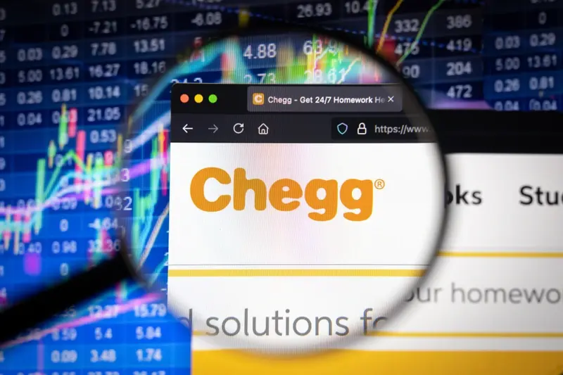 Chegg company logo on a website with blurry stock market developments in the background, seen on a computer screen through a magnifying glass