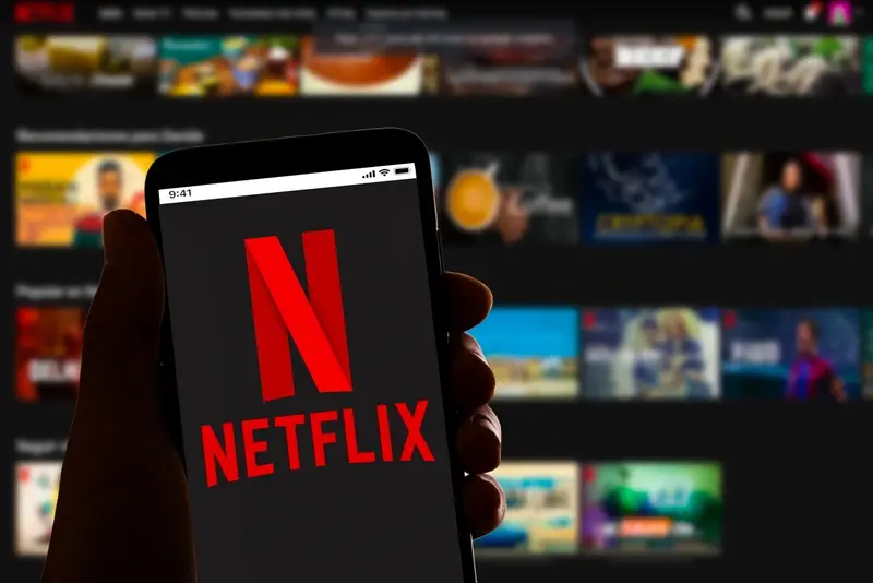 Netflix logo on smartphone in front of screen showing main menu for streaming platform