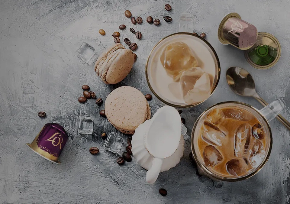 Two iced coffees featuring L'OR Espresso, assorted macarons, and coffee pods are elegantly arranged alongside ice cubes on a textured surface.