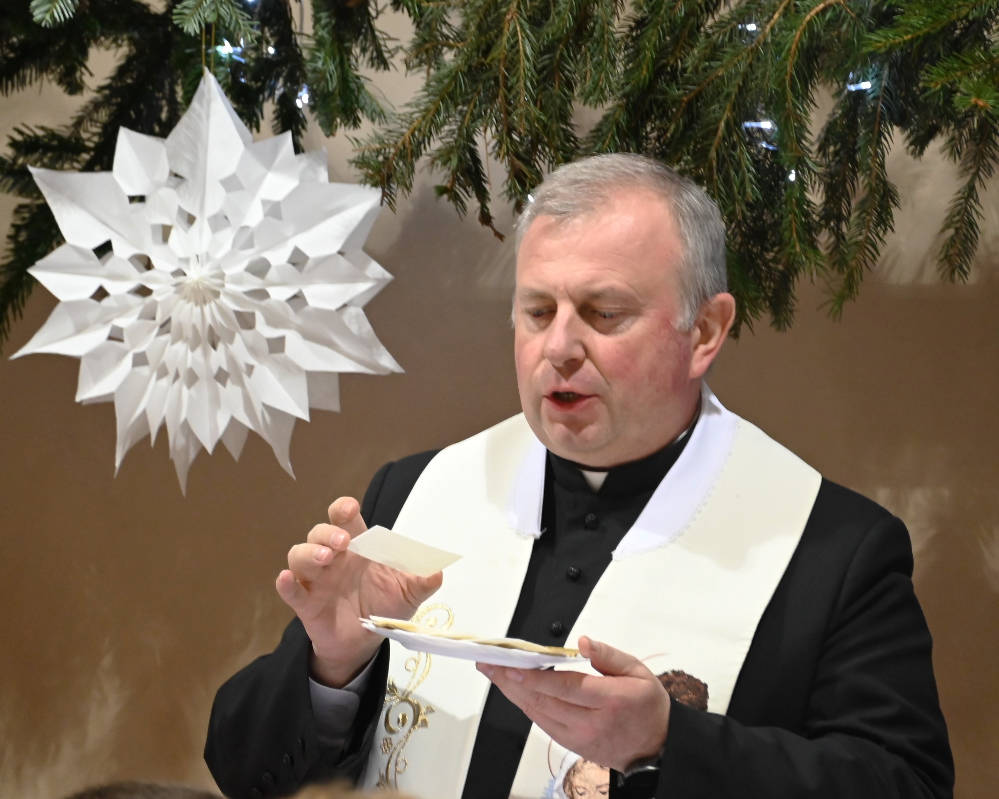 Wigilia w Ośrodku | Ksiądz dziekan trzyma w ręku talerzyk z połatkami, w drugiej ręce unosi jeden z opłatków. W tle dekoracja z gałązek świerka i duża, biała gwiazda z papieru.JPG