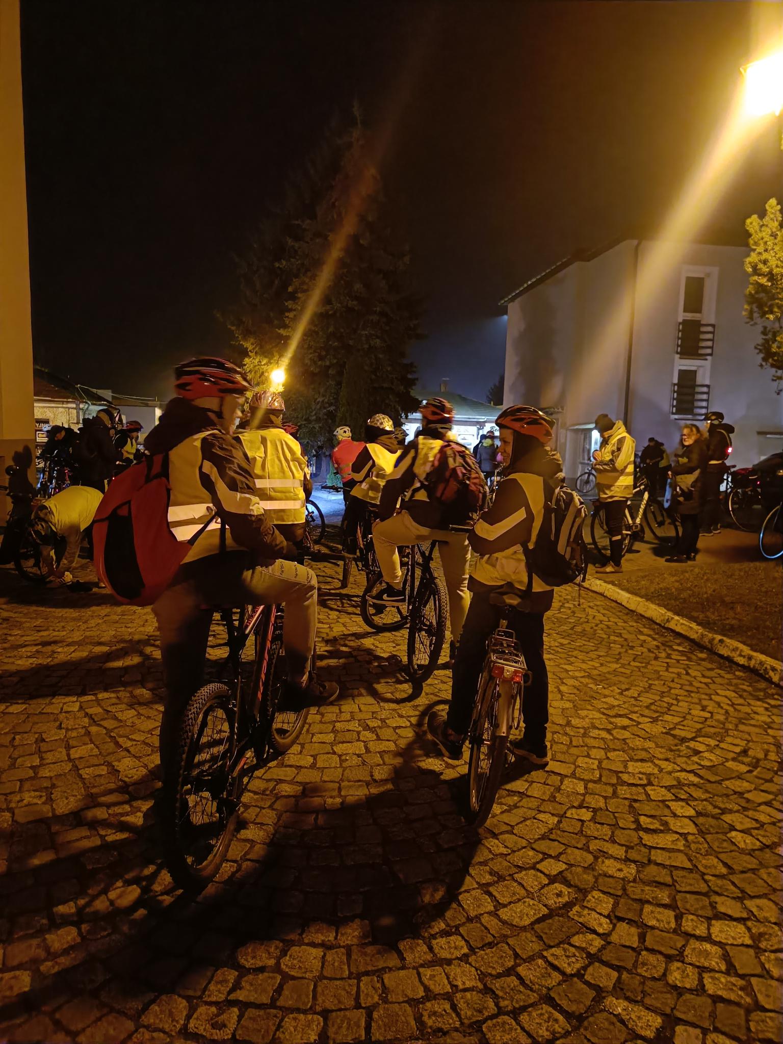 Rowerowa Droga Krzyżowa | Na brukowanym placu siedzą na rowerach wychowankowie MOW ubrani w kaski i kamizelki odblaskowe. Zdjęcie nocne.jpg
