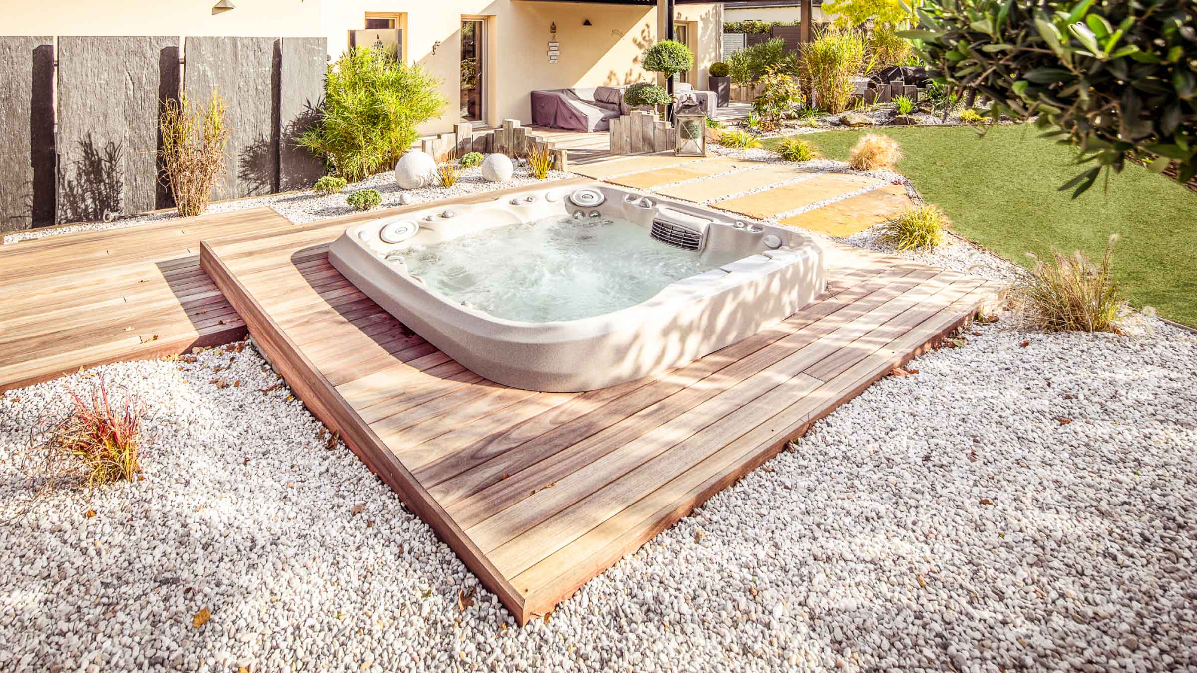 Spa with jets turned on, surrounded in timber decking sits beside a home with green shrubs dotted around.