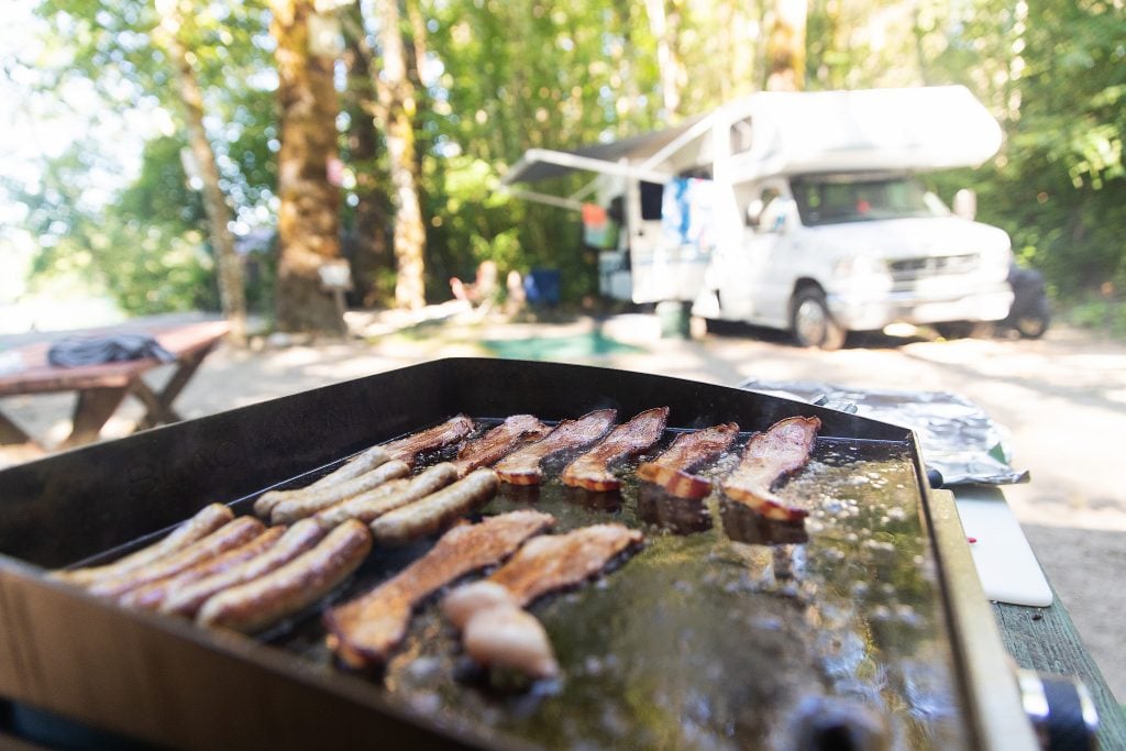 A griddle with bacon and sausage cooking in front of an RV, a great setup for camping meals