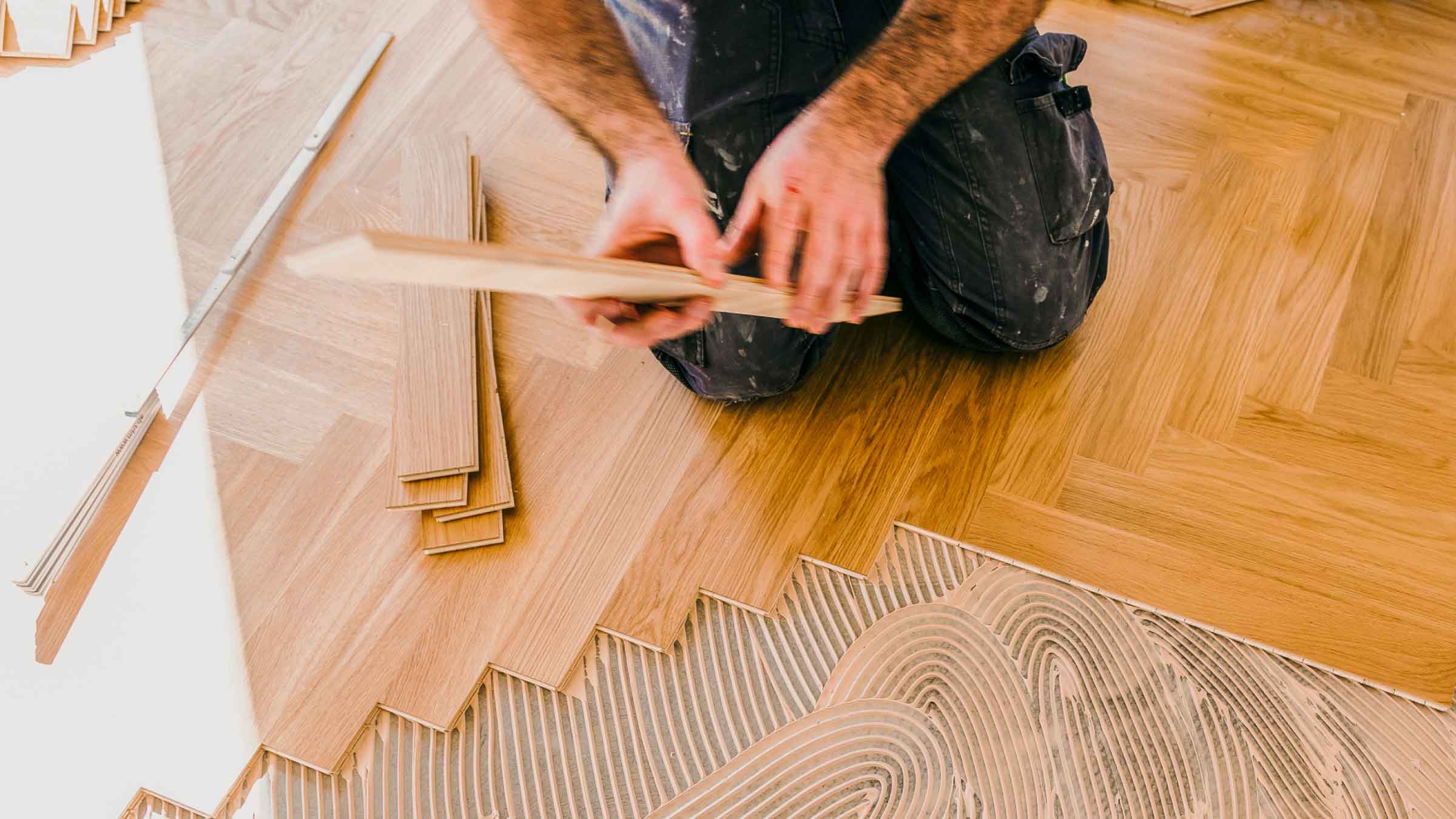 Tradesman in overalls kneels down placing a piece of timber onto a concrete floor where he's almost finished laying a herringbone patterned timber floor.